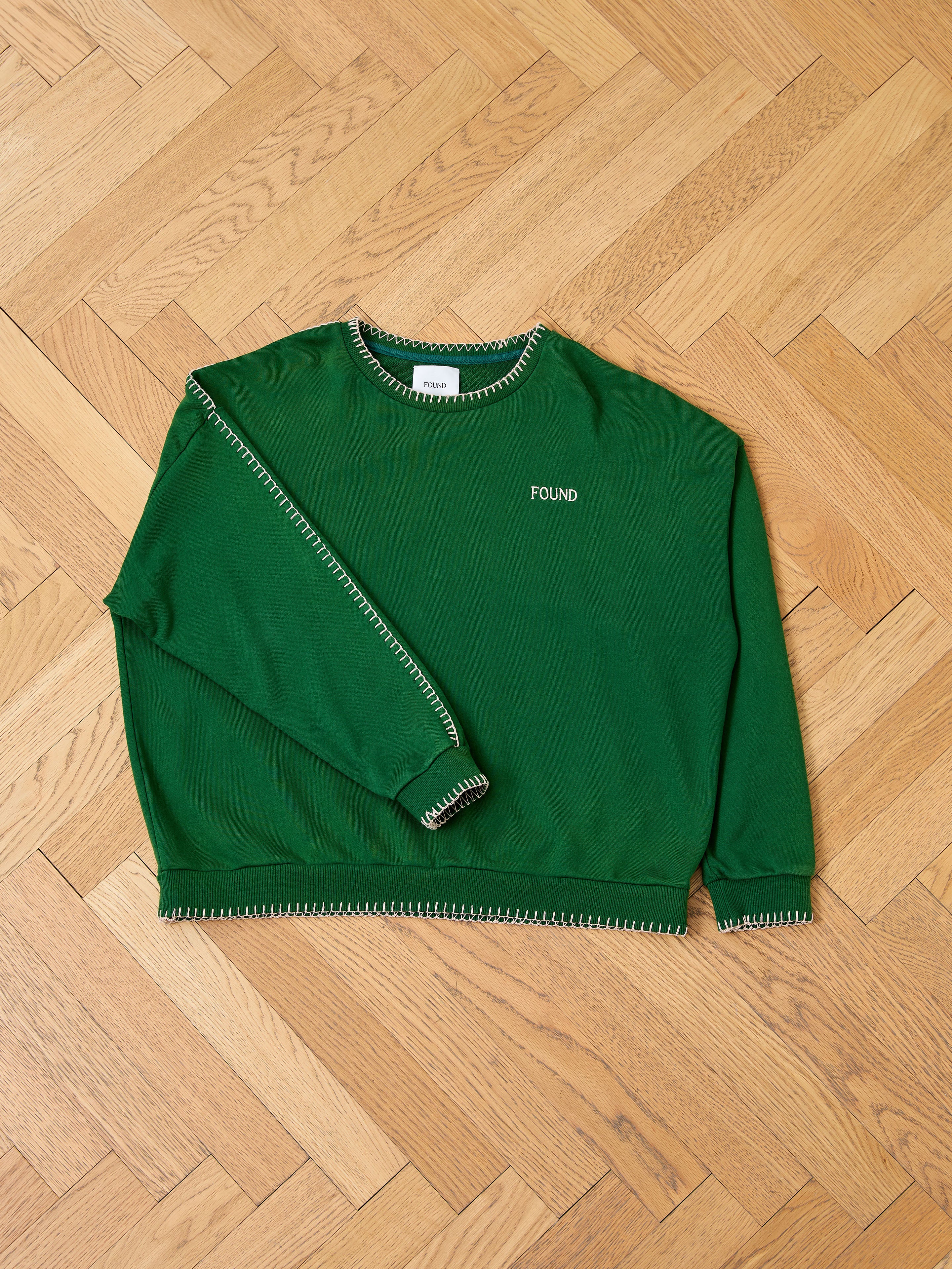 The FOUND Stitched Border Distressed Crewneck, a vintage-style green sweater made from soft French terry cotton, is displayed on a wooden floor.