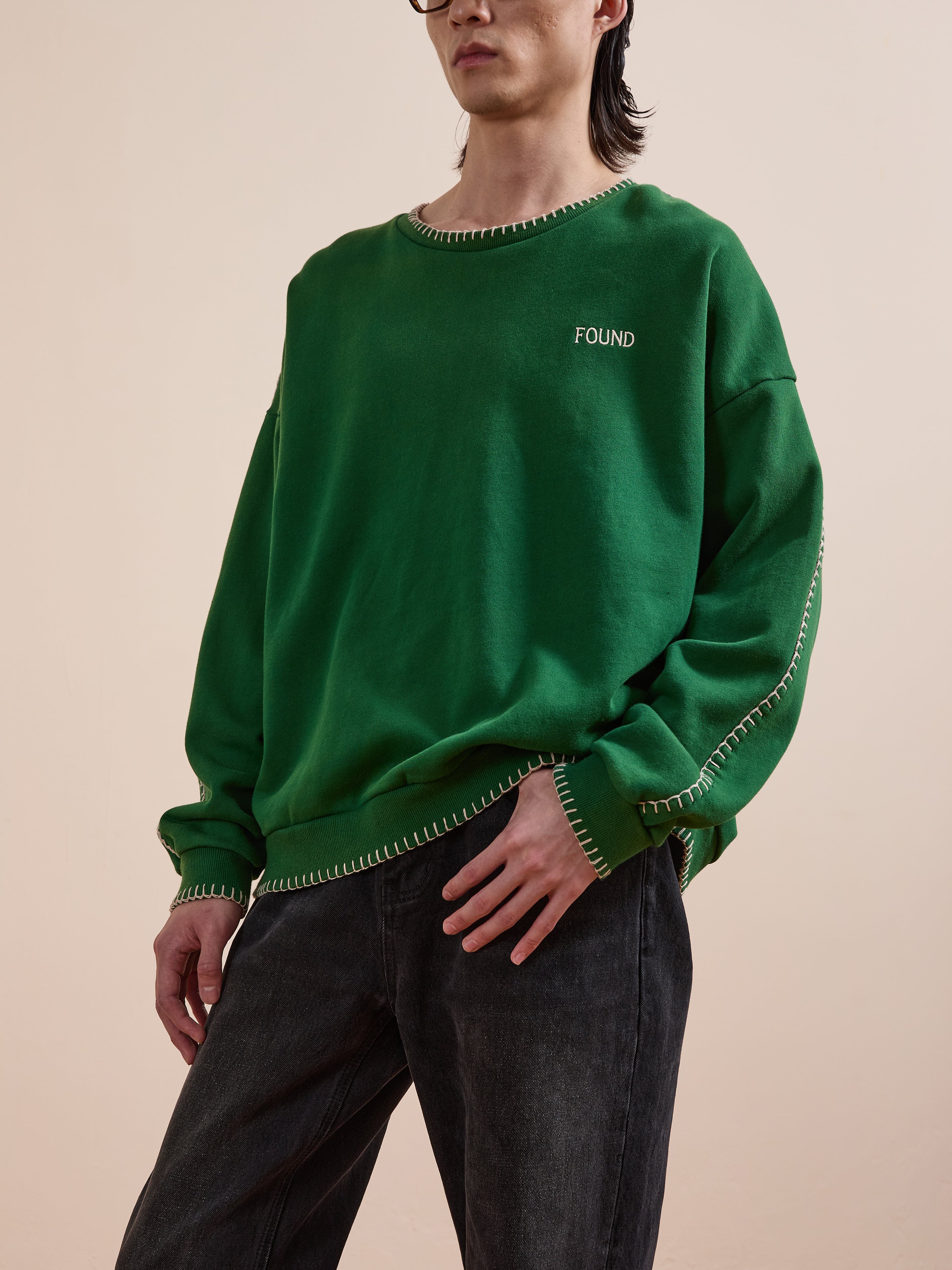 A person wears FOUND’s Stitched Border Distressed Crewneck, a loose green French terry sweatshirt with white stitching and chest embroidery, paired with dark jeans against a plain background.