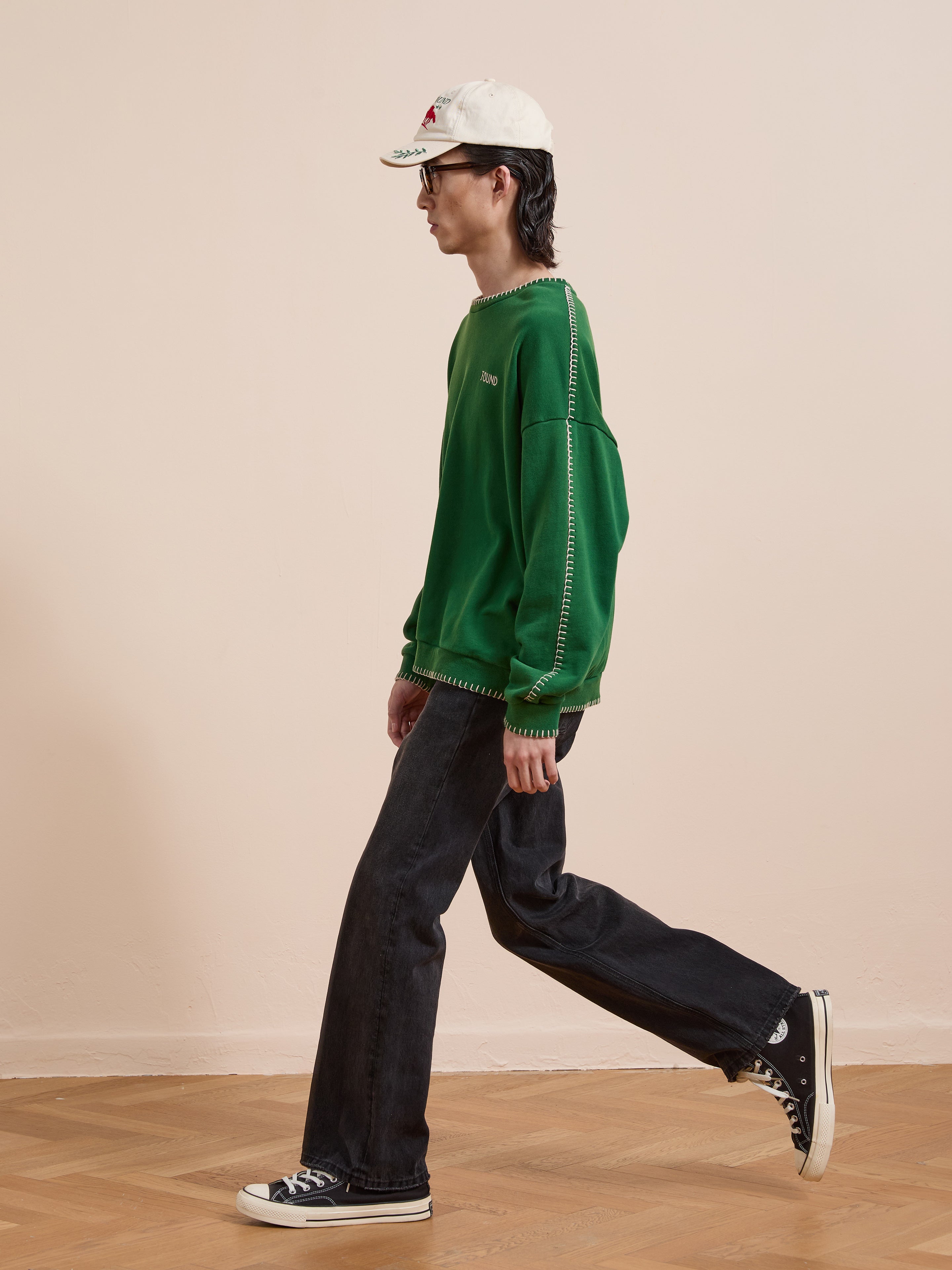 Someone wears the FOUND Stitched Border Distressed Crewneck in green, paired with black jeans, a white cap, and black sneakers, walking indoors on a wooden floor against a plain beige wall.
