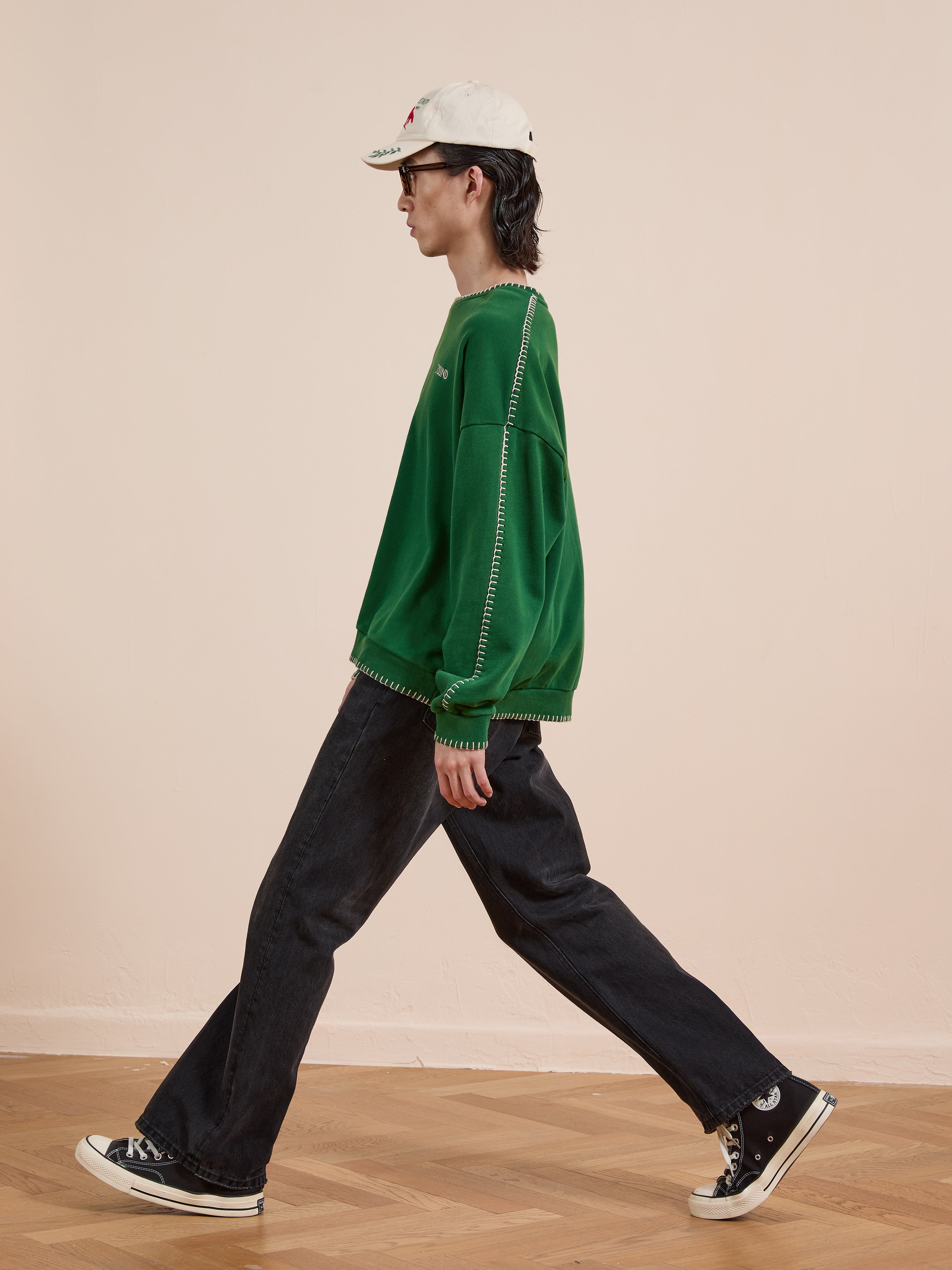 Person in a FOUND Stitched Border Distressed Crewneck, black wide-leg jeans, black Converse sneakers, and a white cap walks indoors on a wooden floor against a plain beige wall.