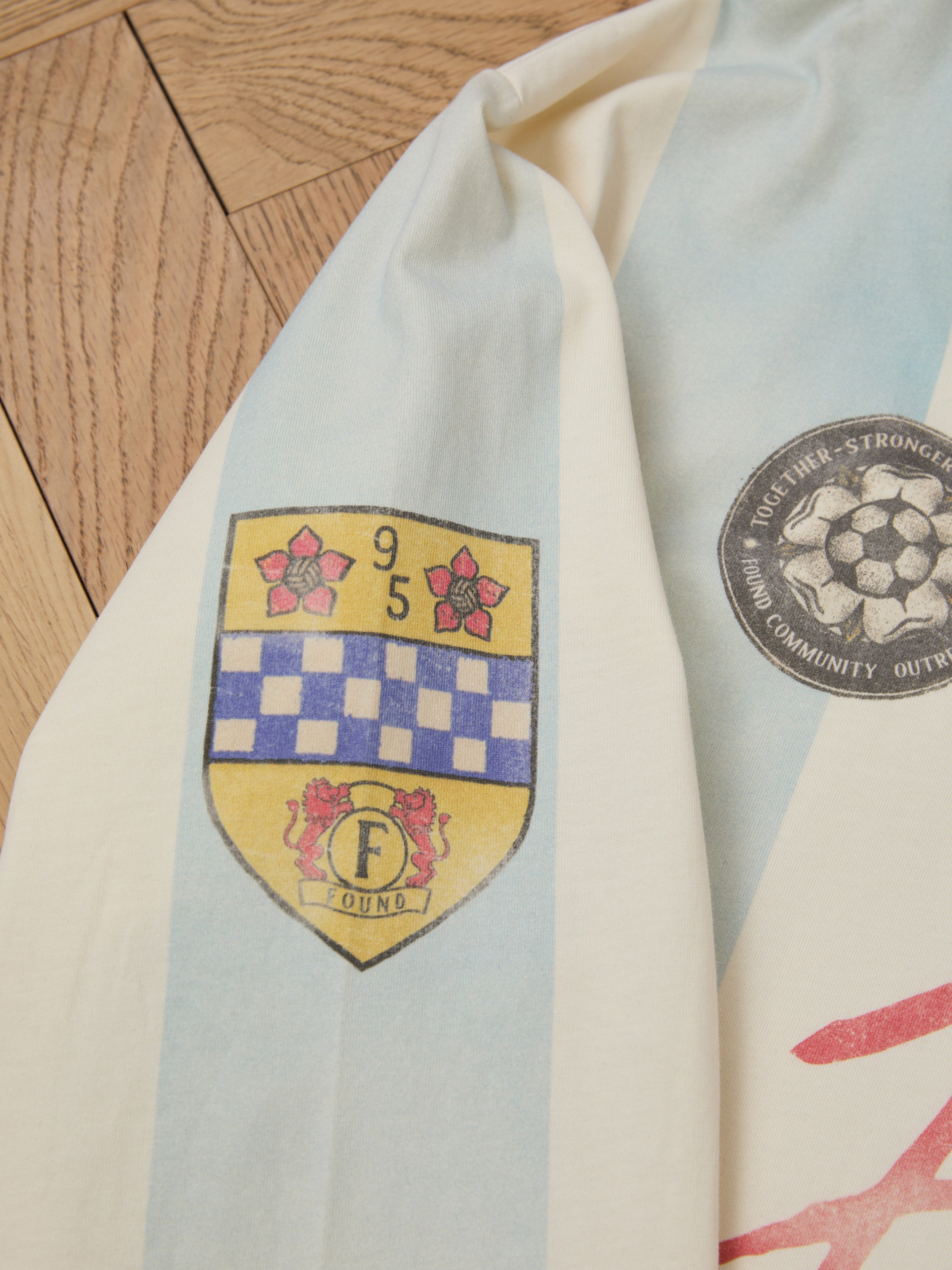 Close-up of the Stripe Sport LS Jersey by FOUND on a wooden floor, featuring a heritage-inspired yellow and blue crest with floral motifs and the number 95, plus a round soccer ball emblem. Its unisex relaxed fit is ideal for sports enthusiasts seeking style and comfort.