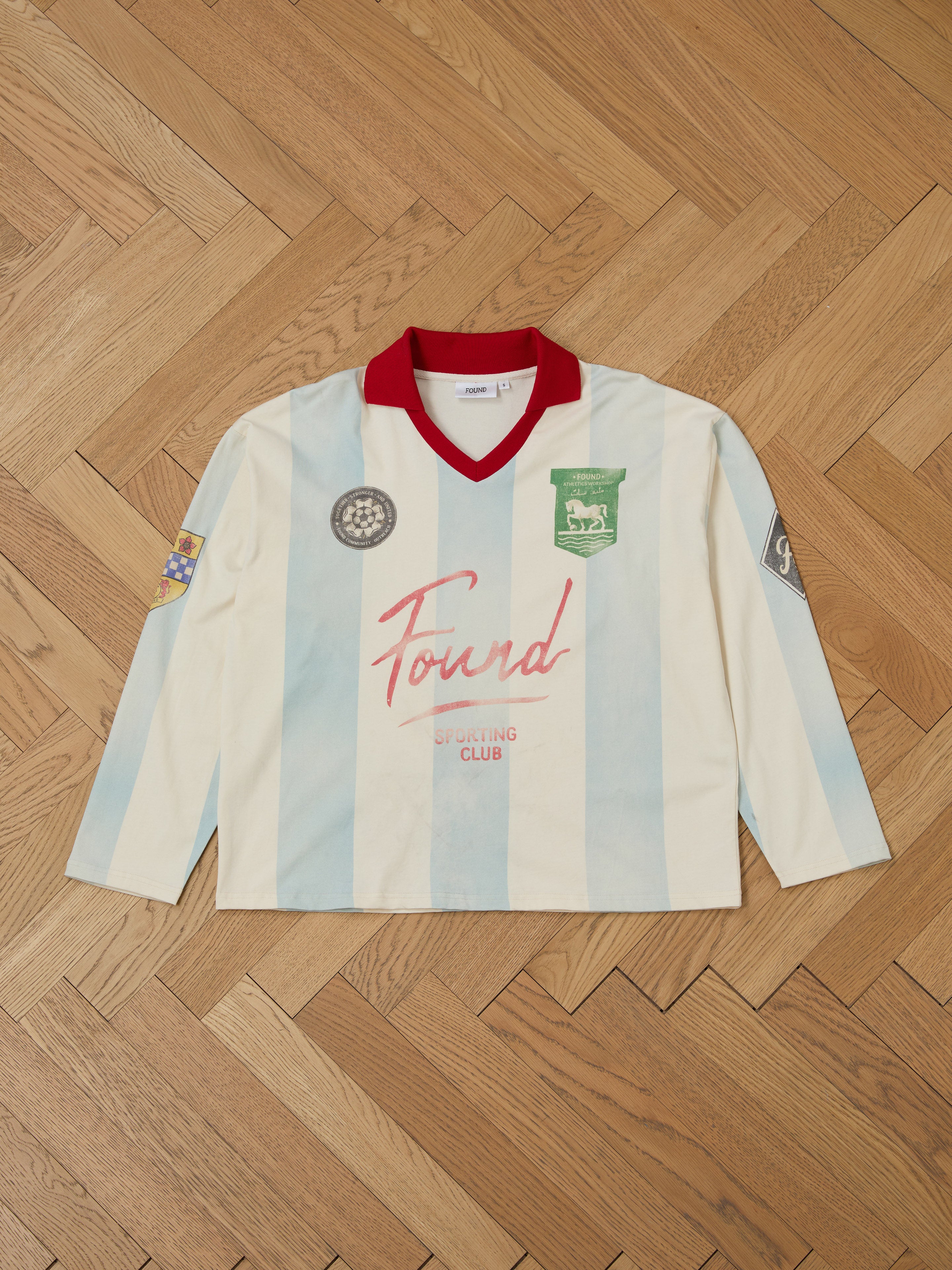 The Striped Sport LS Jersey by FOUND is a vintage sportswear piece with light blue and white stripes, a red collar, and placed on a wooden floor. It features green and black emblems on the left chest along with "FOUND" logos.