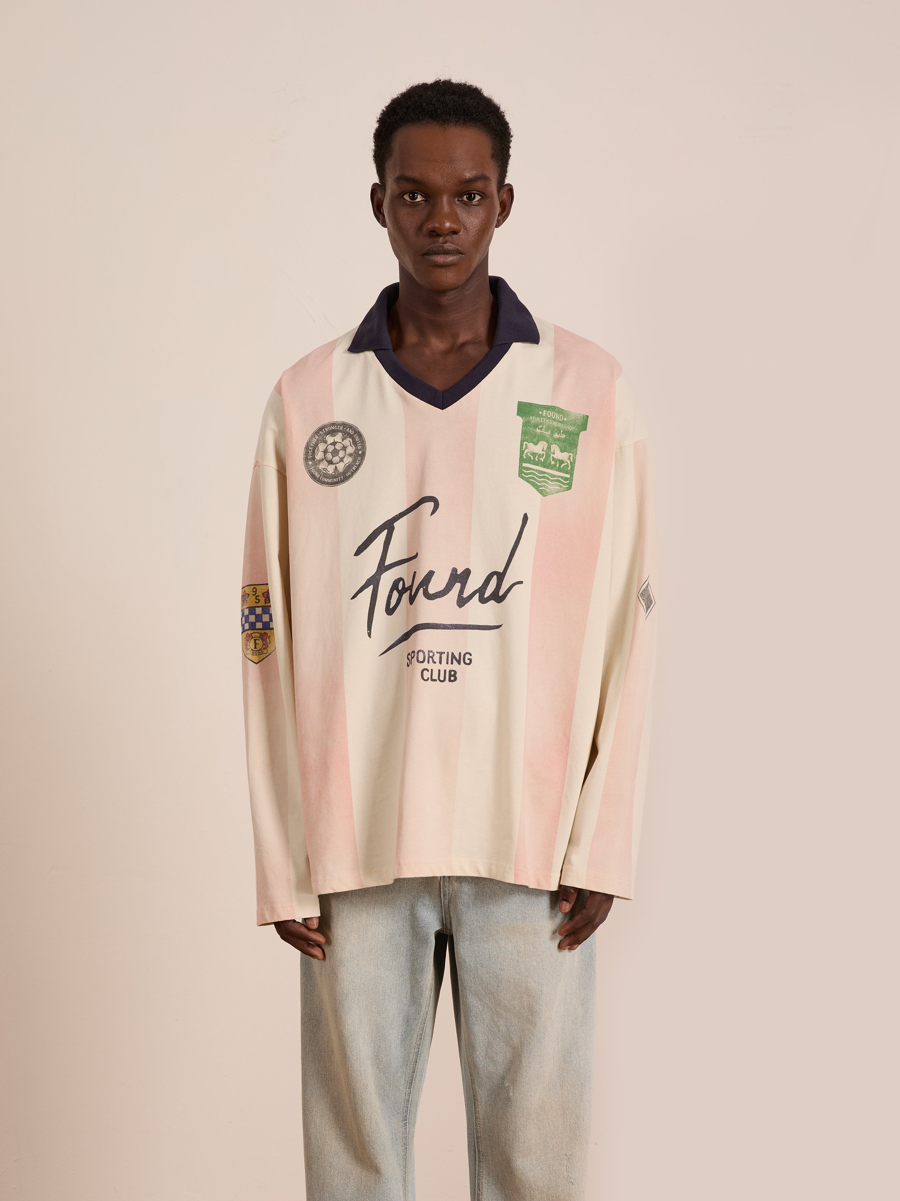 Someone stands against a plain background wearing the FOUND Striped Sport LS Jersey— a unisex, relaxed-fit, long-sleeve cream and pink top with "Found Sporting Club" text and patches—paired with light blue jeans.