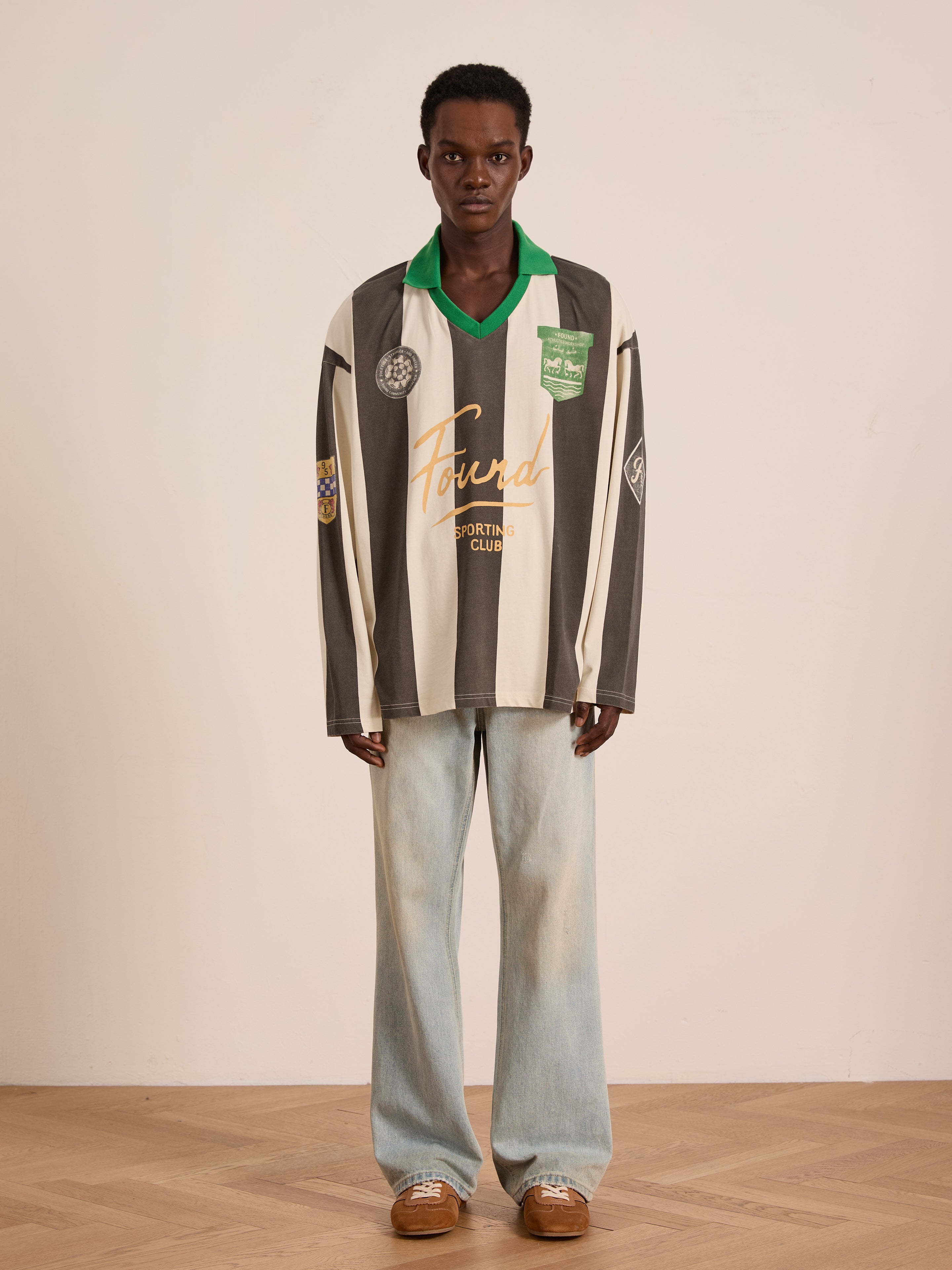 A person stands indoors against a light wall wearing the FOUND Striped Sport LS Jersey in black-and-white stripes, paired with light blue wide-leg jeans and brown shoes. The relaxed fit jersey is unisex.