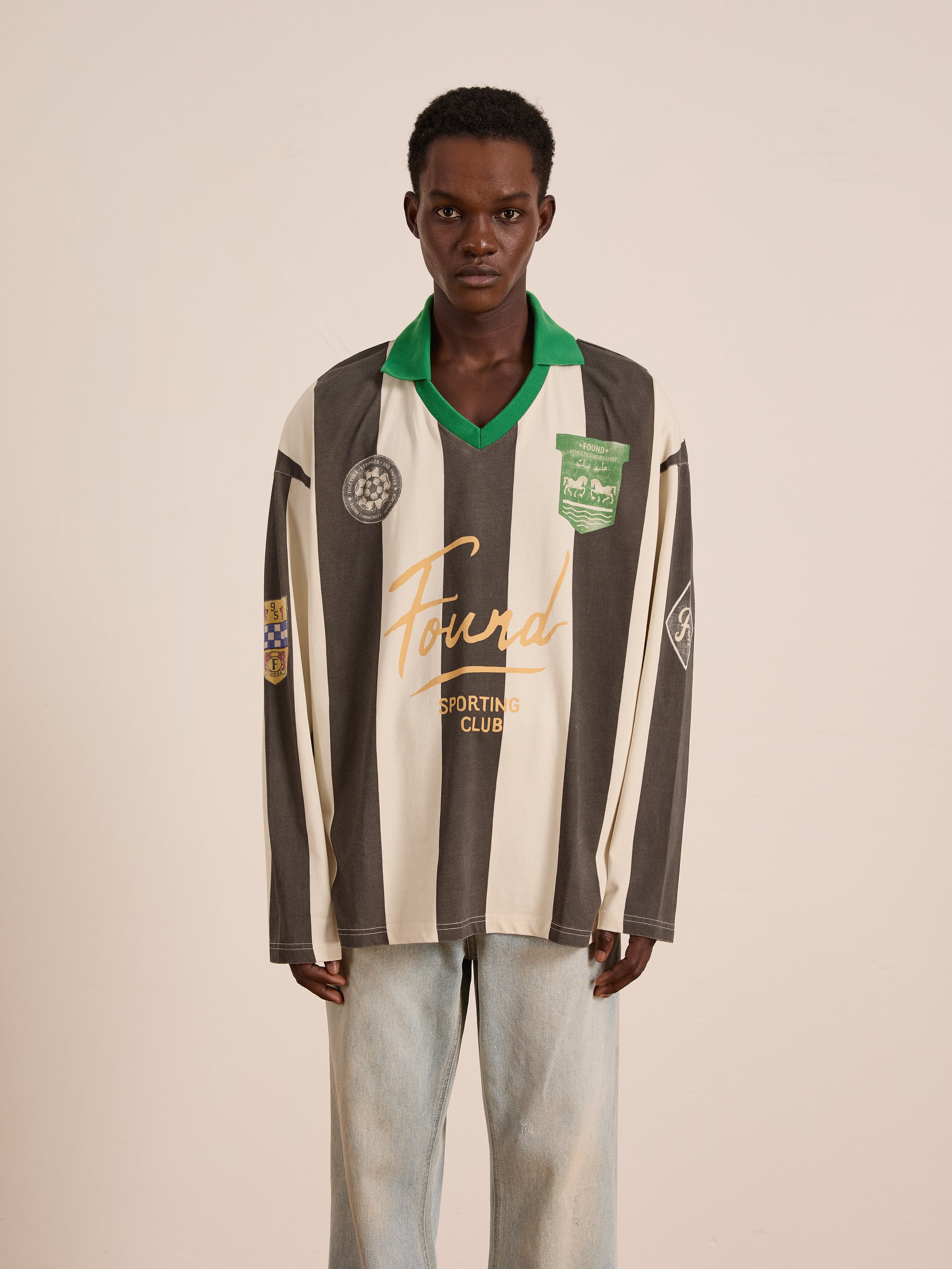 A person wearing the FOUND Striped Sport LS Jersey—a relaxed fit, unisex black-and-white striped top with logos—paired with light blue jeans stands against a plain beige background.