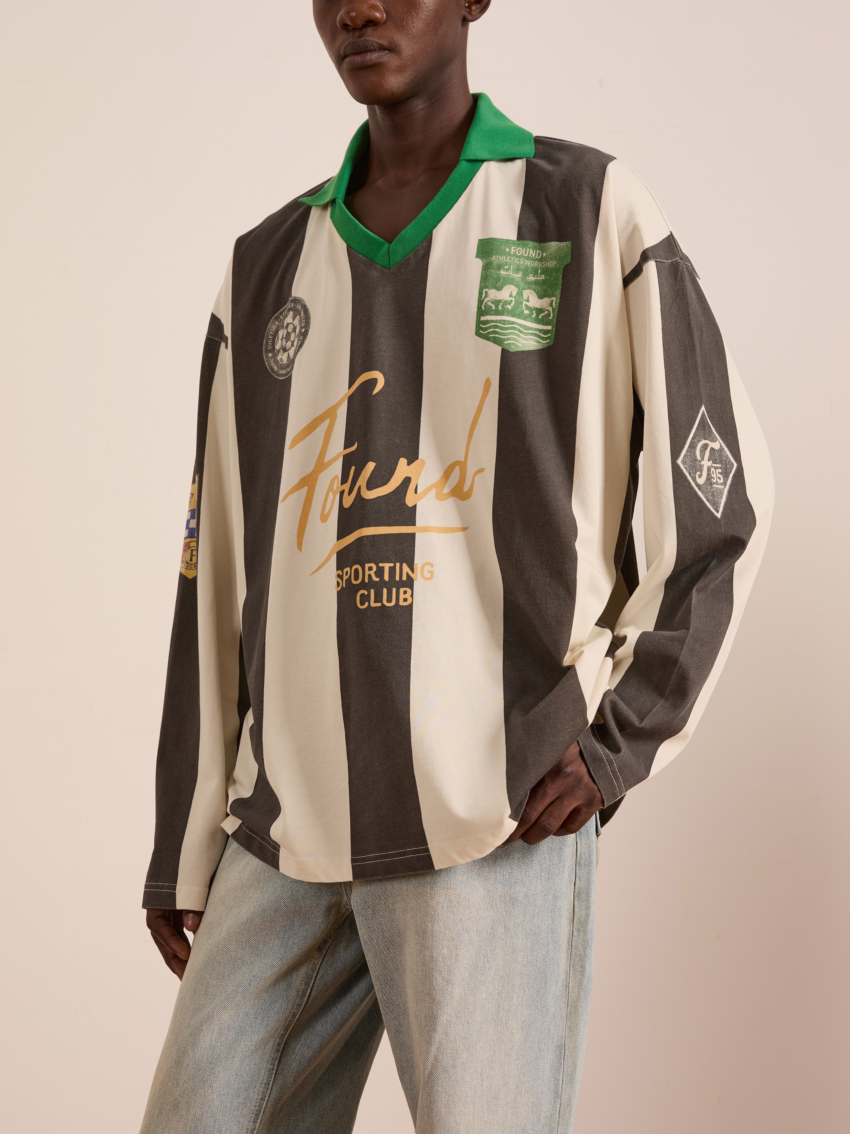A person wears the FOUND Striped Sport LS Jersey, a unisex long-sleeved top with green collar, patches, and "Found Sporting Club" logo, styled with light blue relaxed-fit jeans.
