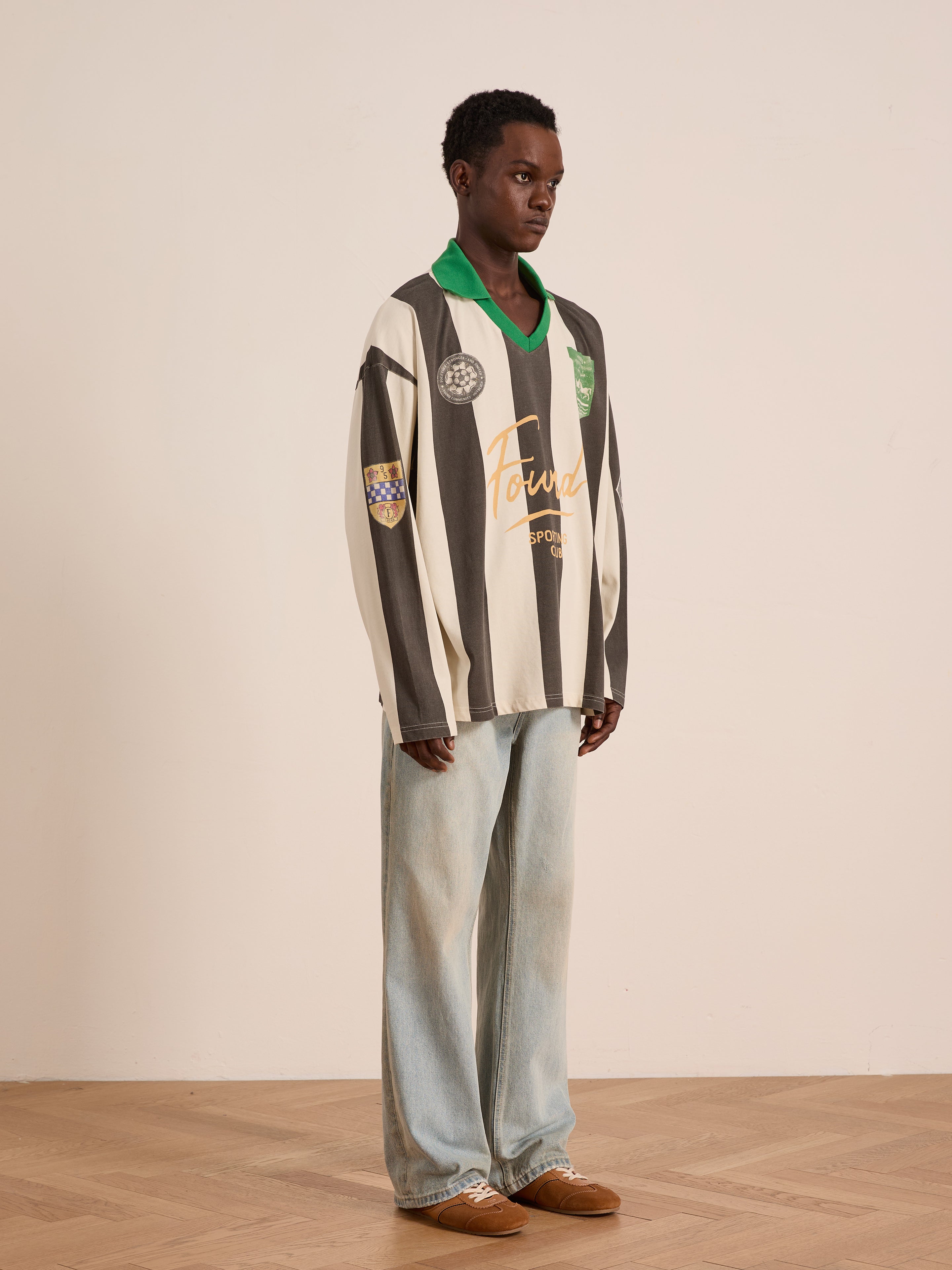 Indoors against a beige wall, a person wears the FOUND Striped Sport LS Jersey in black and white, styled with loose light blue jeans and brown shoes.