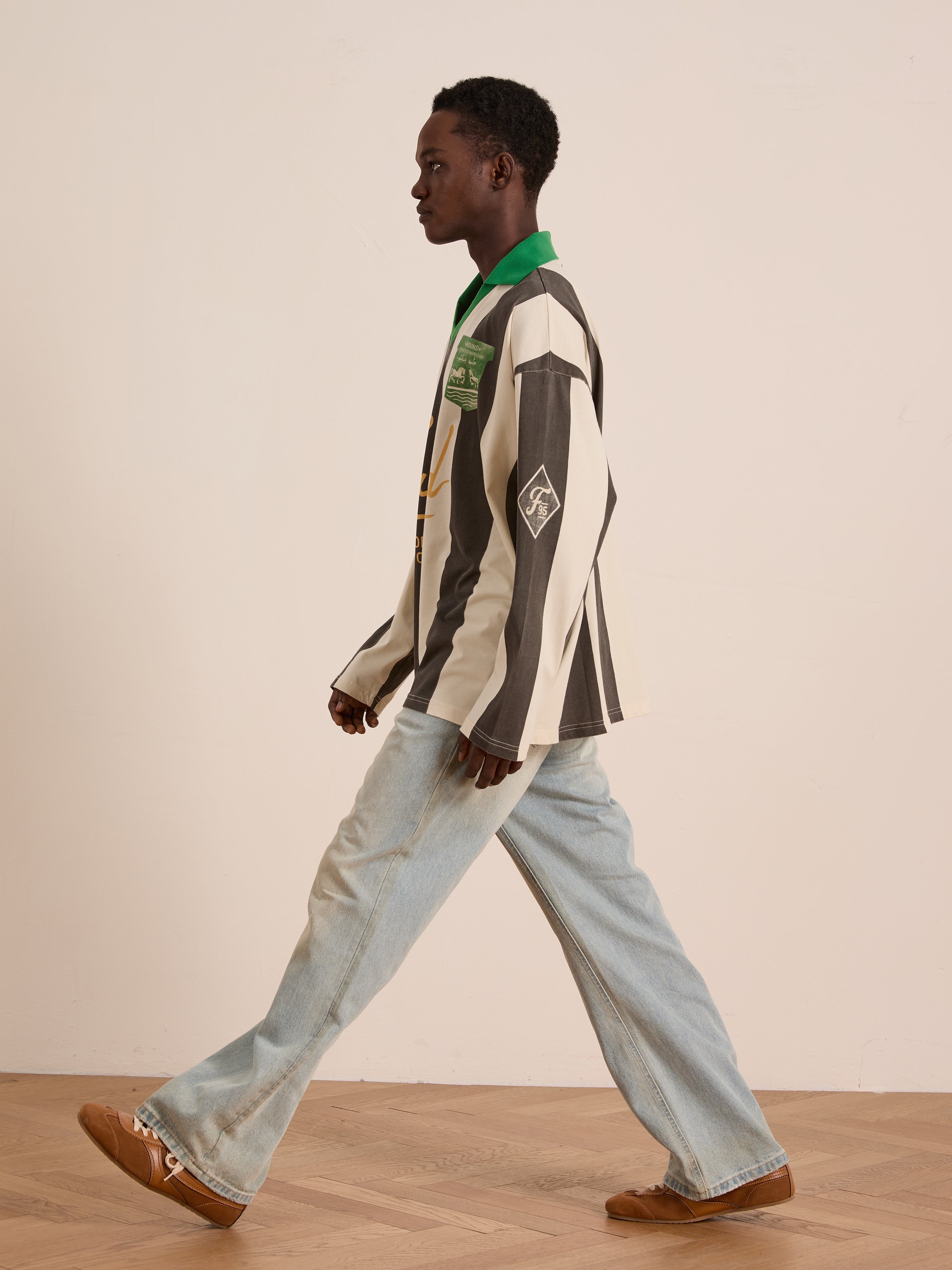 A person in the FOUND Striped Sport LS Jersey, light blue jeans, and brown shoes walks indoors on a wooden floor against a plain wall.