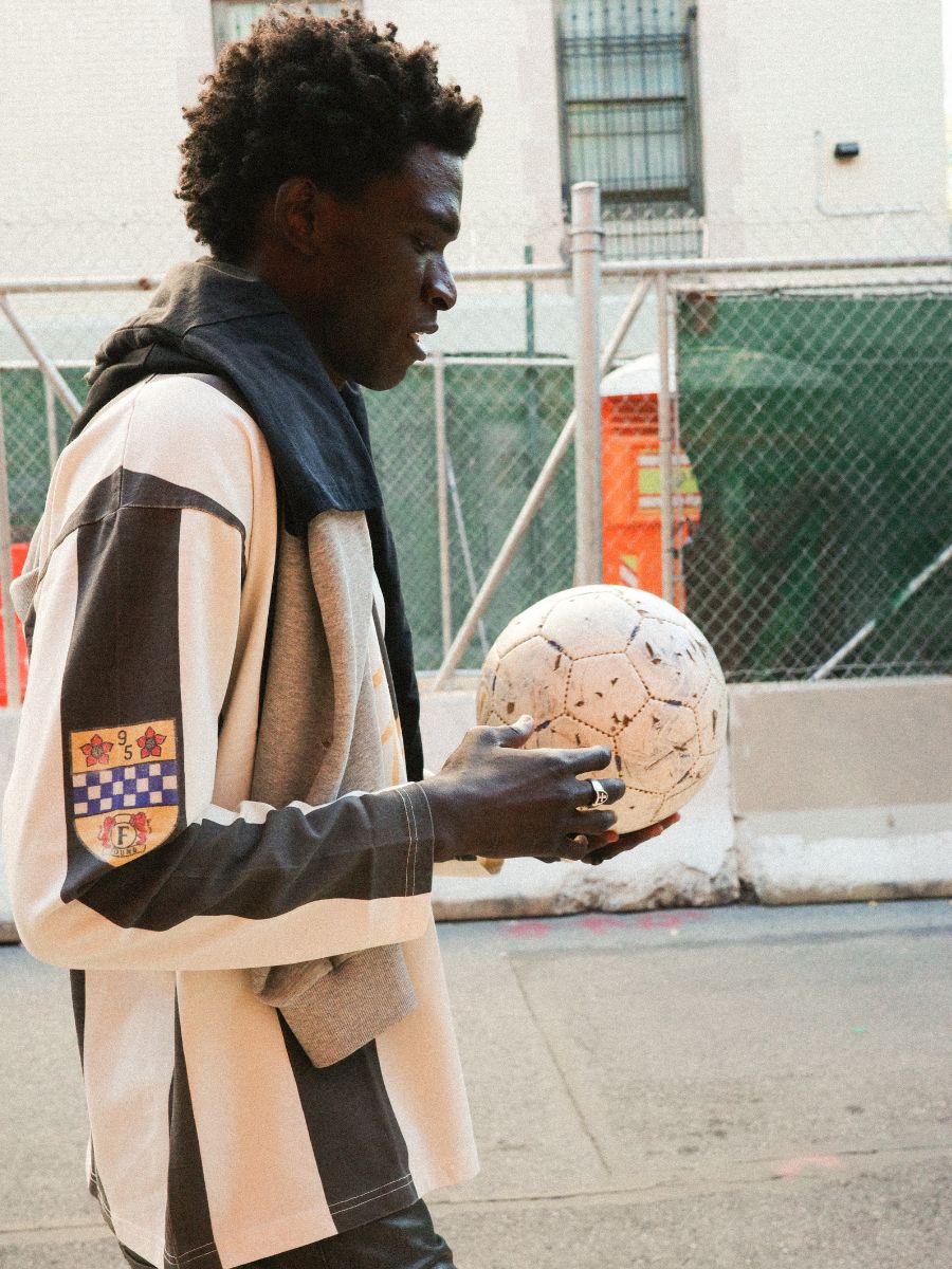 Wearing the FOUND Striped Sport LS Jersey with a jacket draped over their shoulders, a person holds a soccer ball while walking near a city construction site.