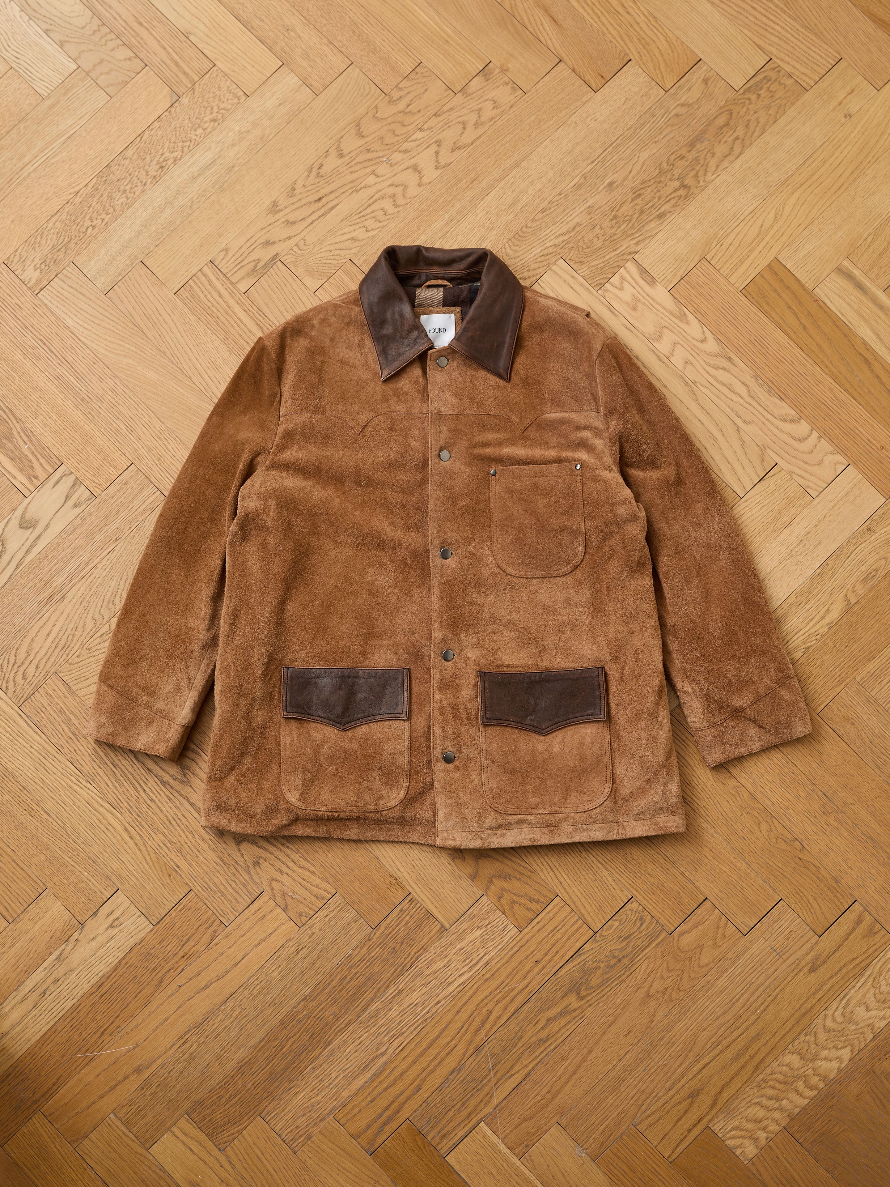 The FOUND Suede Contrast Chore Jacket features vintage-inspired suede with a dark brown leather collar and pocket accents, shown flat on a wooden herringbone-patterned floor.
