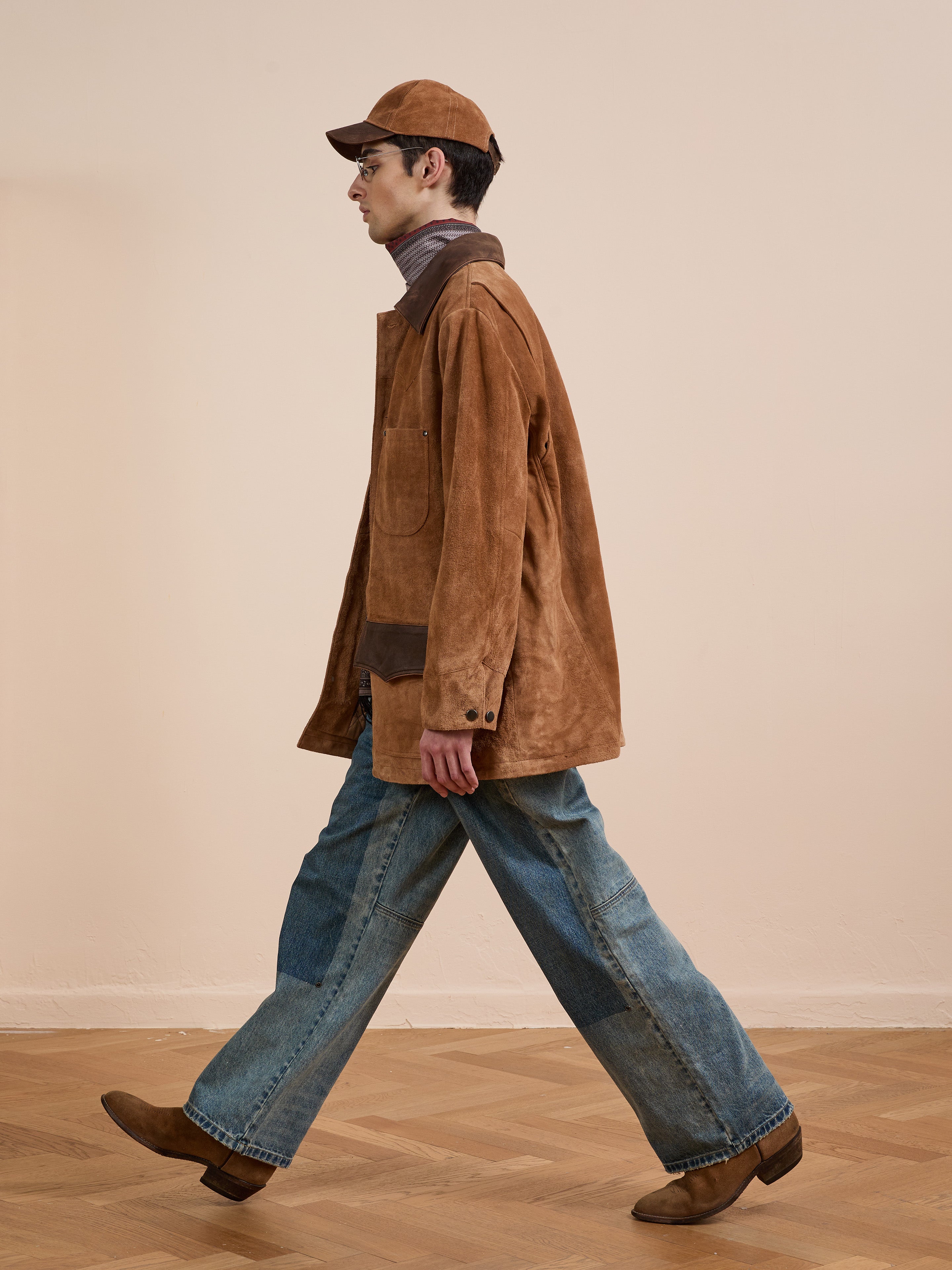 Wearing the FOUND Suede Contrast Chore Jacket, blue jeans, brown boots, and a matching brown cap, a person walks across a wooden floor against a plain beige wall.