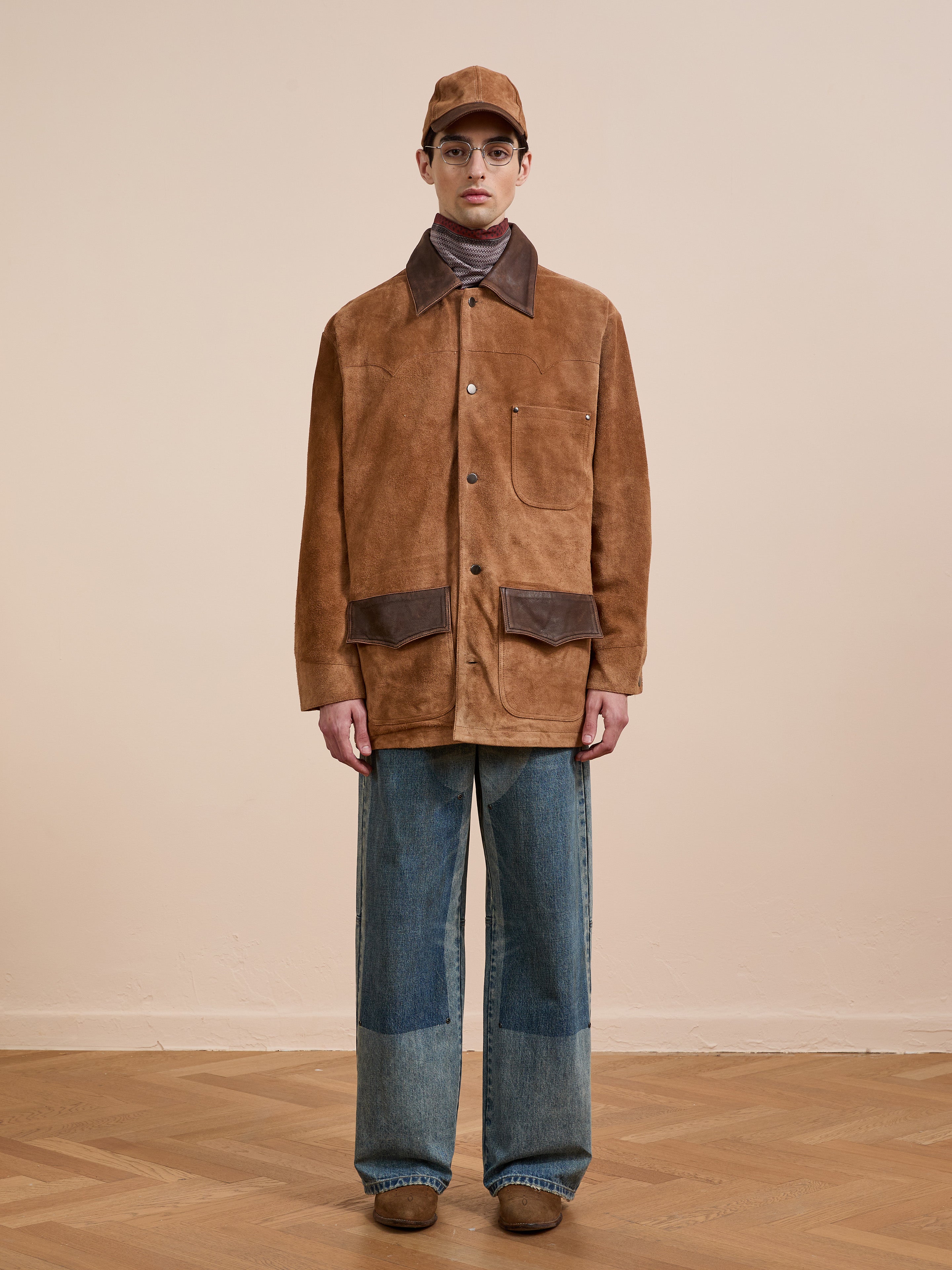 Indoors, a person wears the FOUND Suede Contrast Chore Jacket, blue patched wide-leg jeans, brown shoes, a brown cap, and glasses.