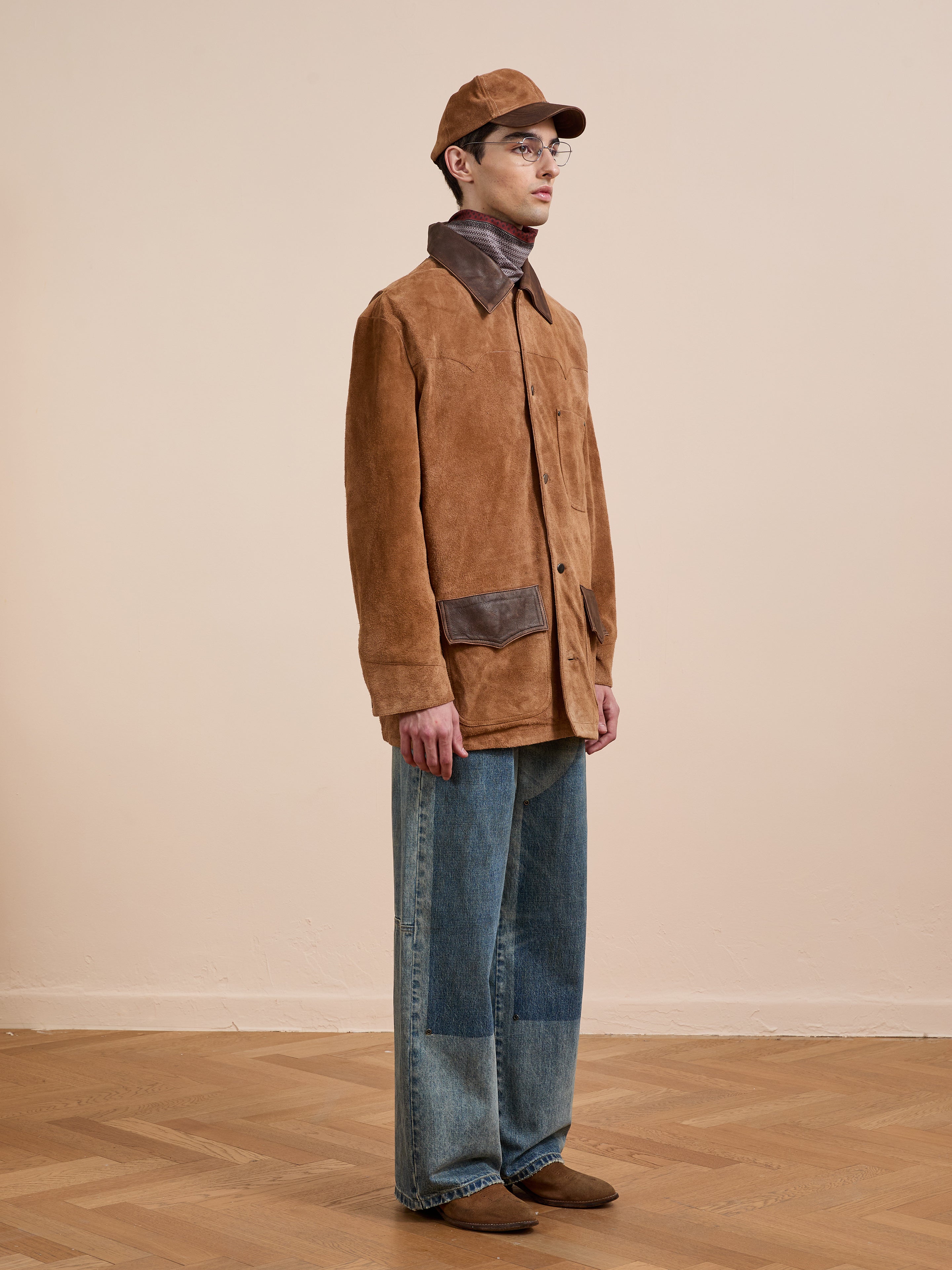 A person in the FOUND Suede Contrast Chore Jacket, featuring a dark brown collar and pocket accents, pairs it with blue patchwork jeans, brown shoes, a brown cap, and glasses while standing on a wooden floor against a beige wall.