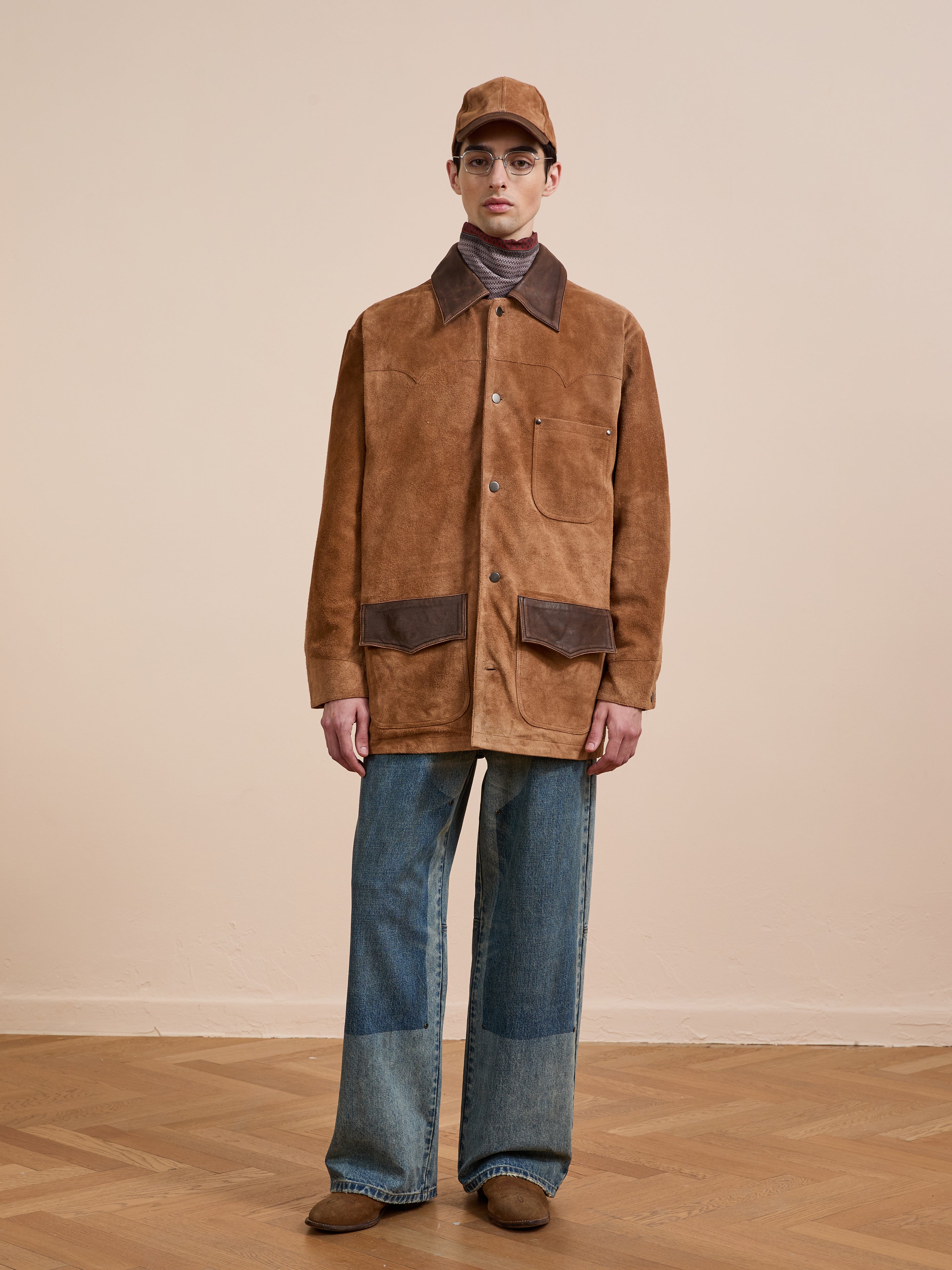 Indoors, a person wears the FOUND Suede Contrast Chore Jacket, paired with blue patchwork jeans, brown shoes, a brown cap, and glasses.