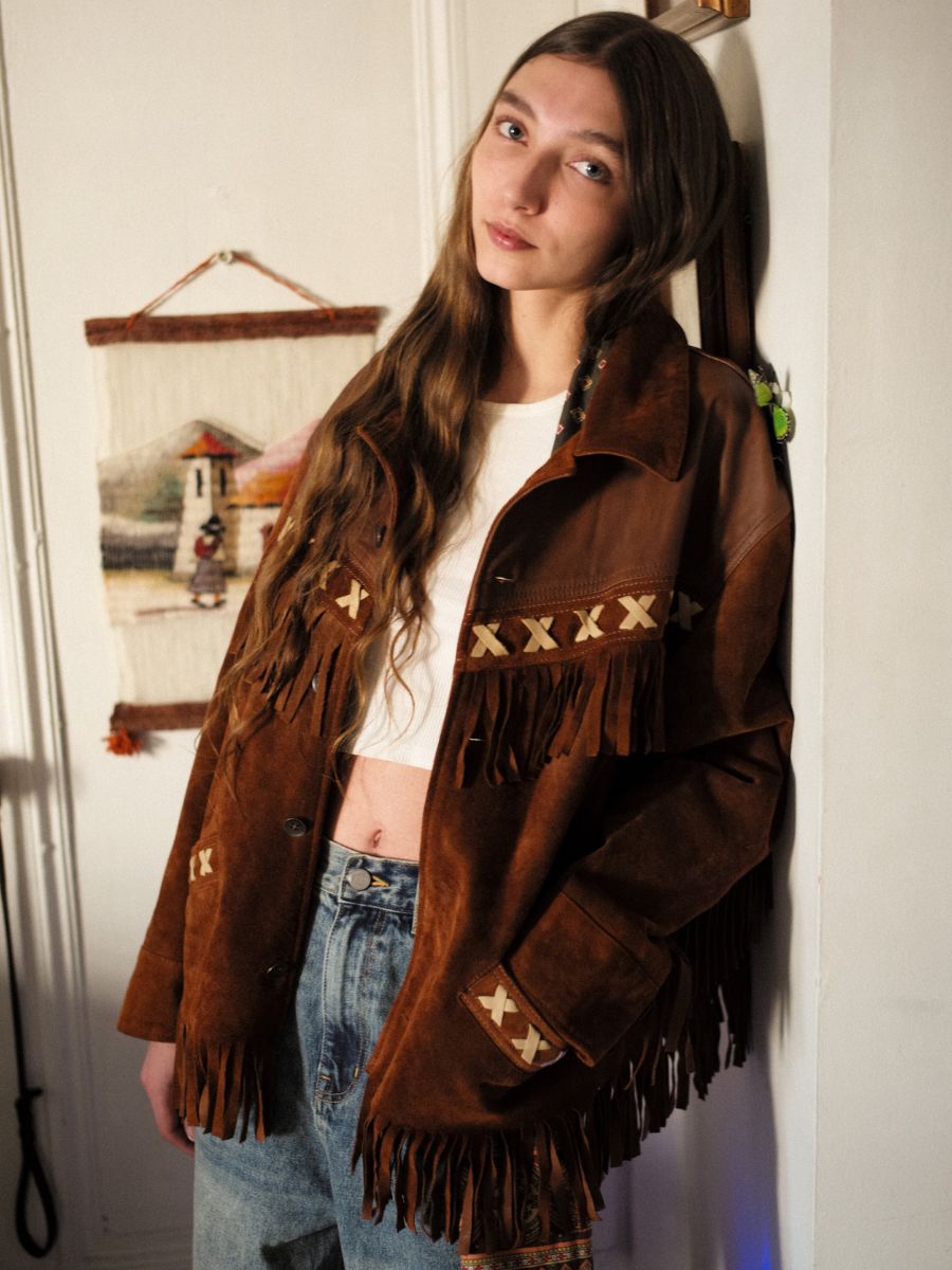 A person with long hair wears a FOUND Western Fringe Suede Leather Jacket, paired with a white top and jeans, standing indoors against a wall featuring an ethically sourced woven hanging.