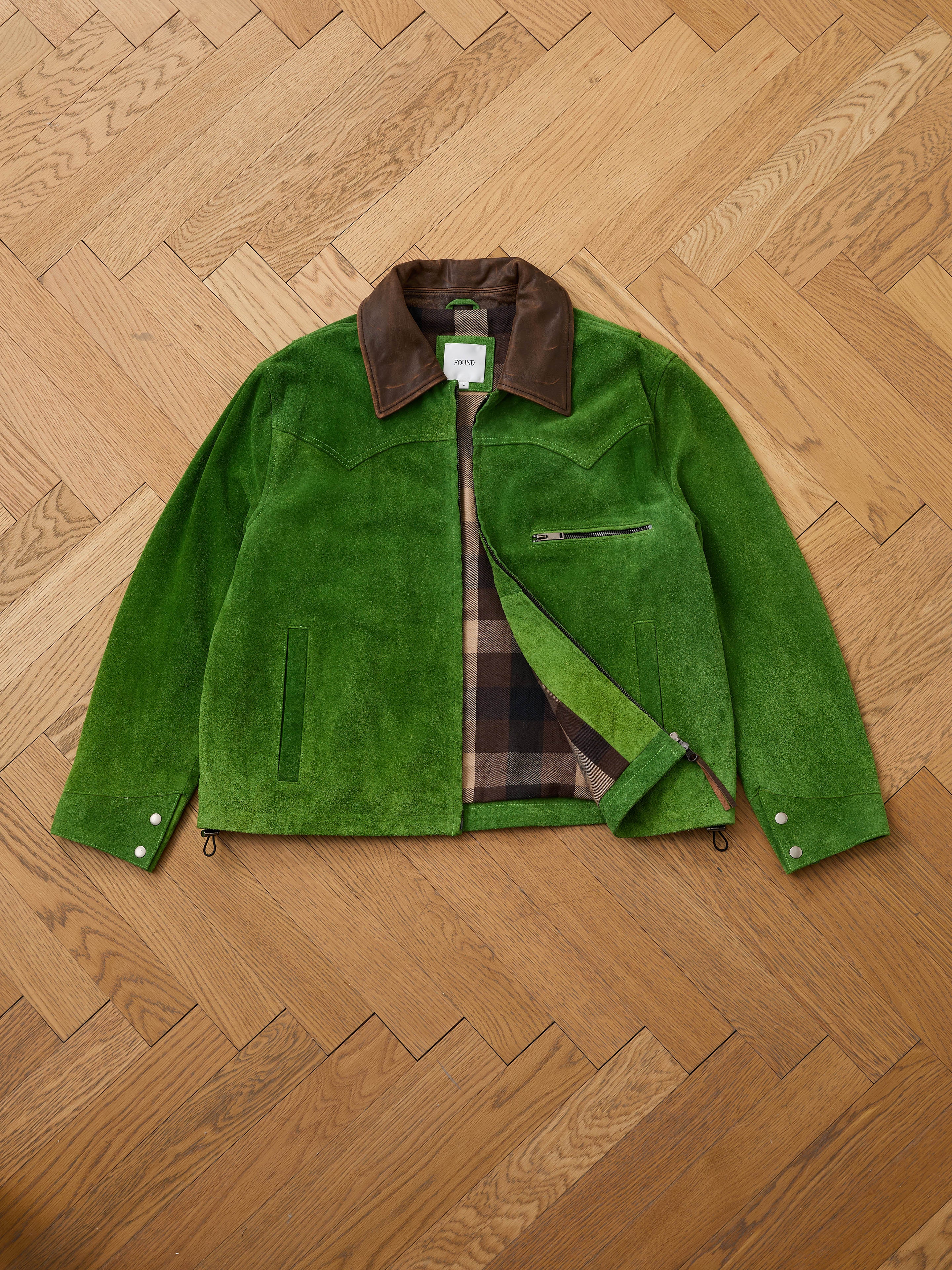 The FOUND Suede Western Barn Jacket features green suede, a brown leather collar, western yoke stitching, zippered chest pocket, and plaid lining, shown laid flat on a wooden herringbone floor.