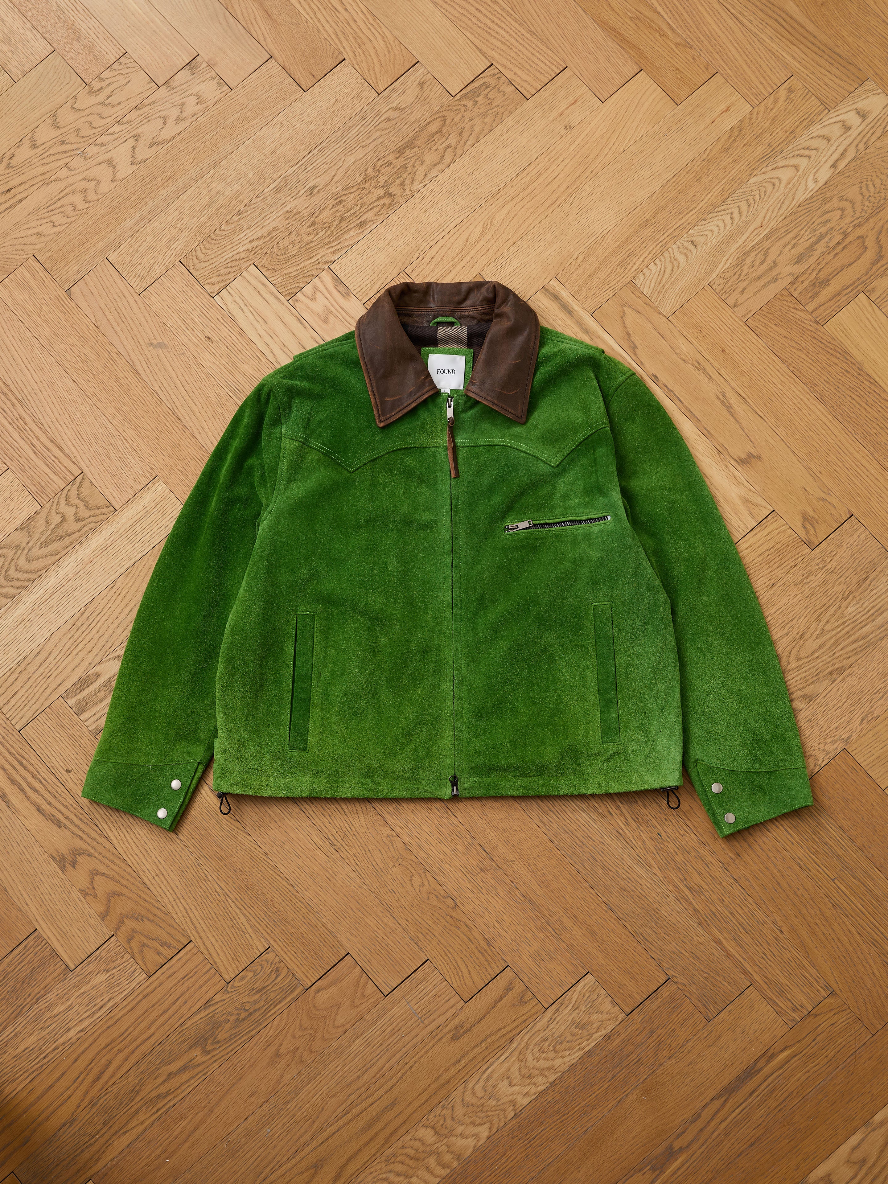 The FOUND Suede Western Barn Jacket in green features a brown genuine suede collar, western yoke stitching, a chest zipper pocket, and buttoned cuffs, shown laid flat on a wooden herringbone floor.