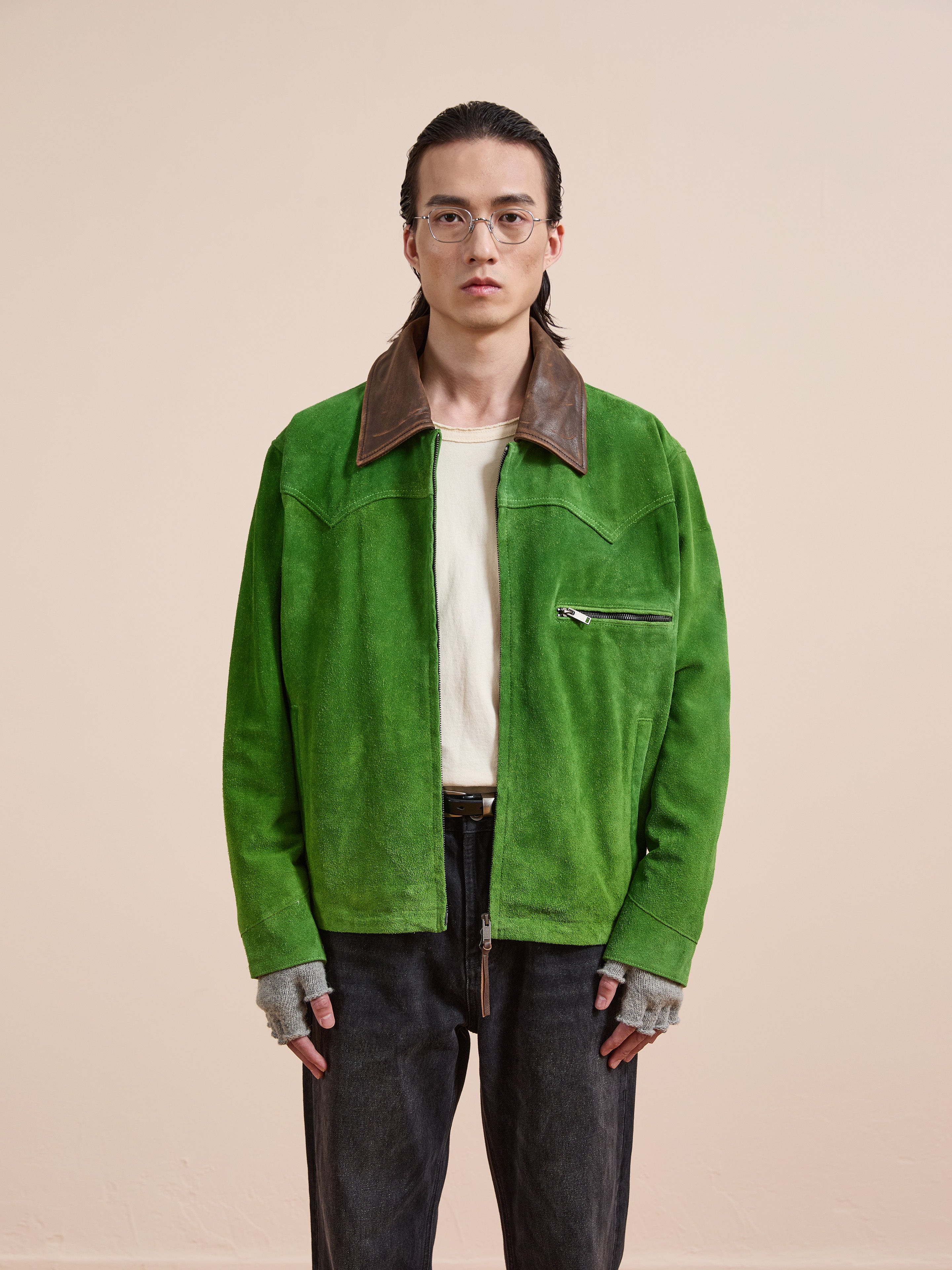 A person with long hair and glasses wears the FOUND Suede Western Barn Jacket, a white shirt, fingerless gloves, and black jeans, standing against a plain beige background.