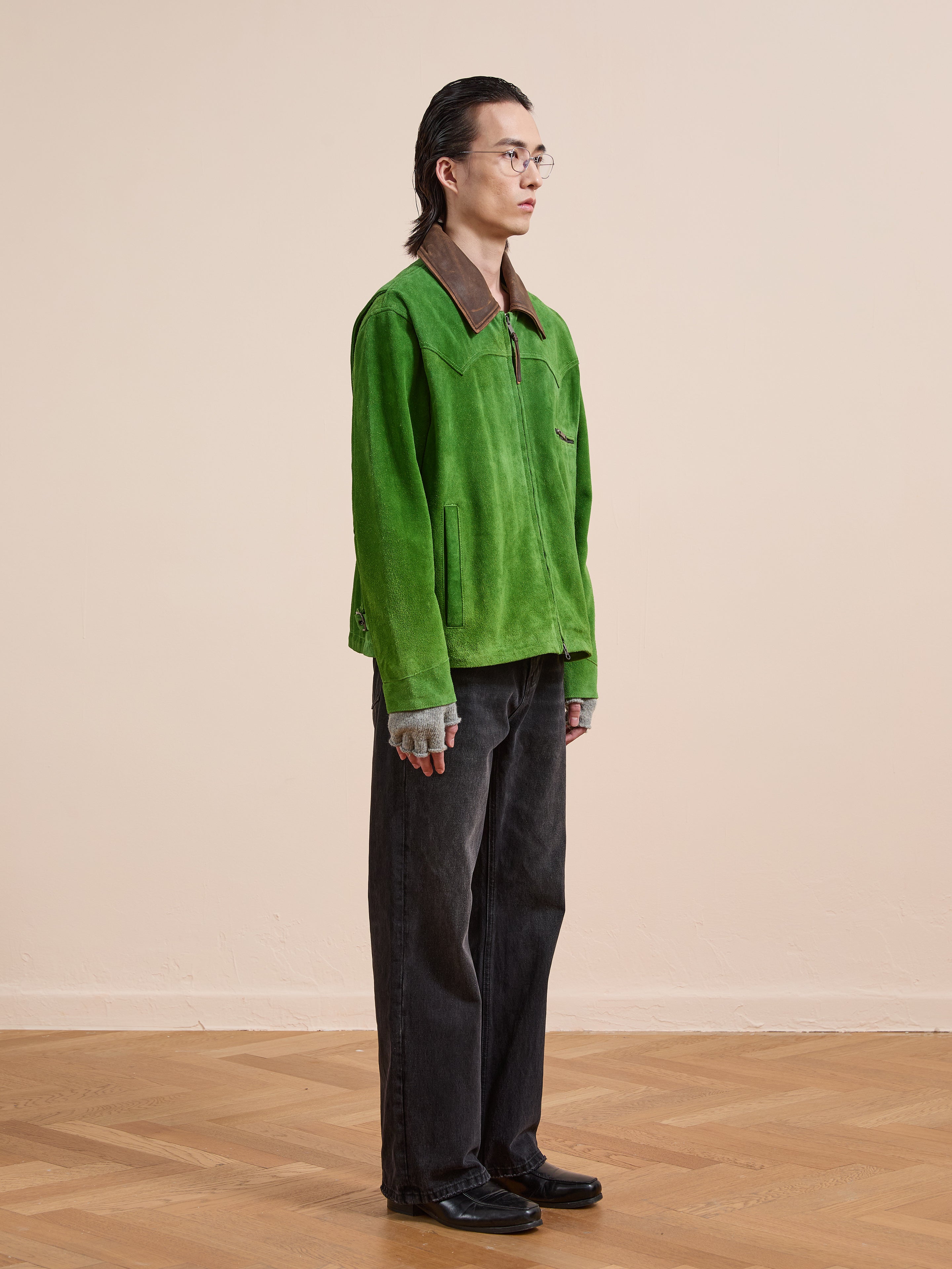 A person stands indoors on a wooden floor, facing right, wearing the FOUND Suede Western Barn Jacket, gray gloves, dark pants, black shoes, and glasses.