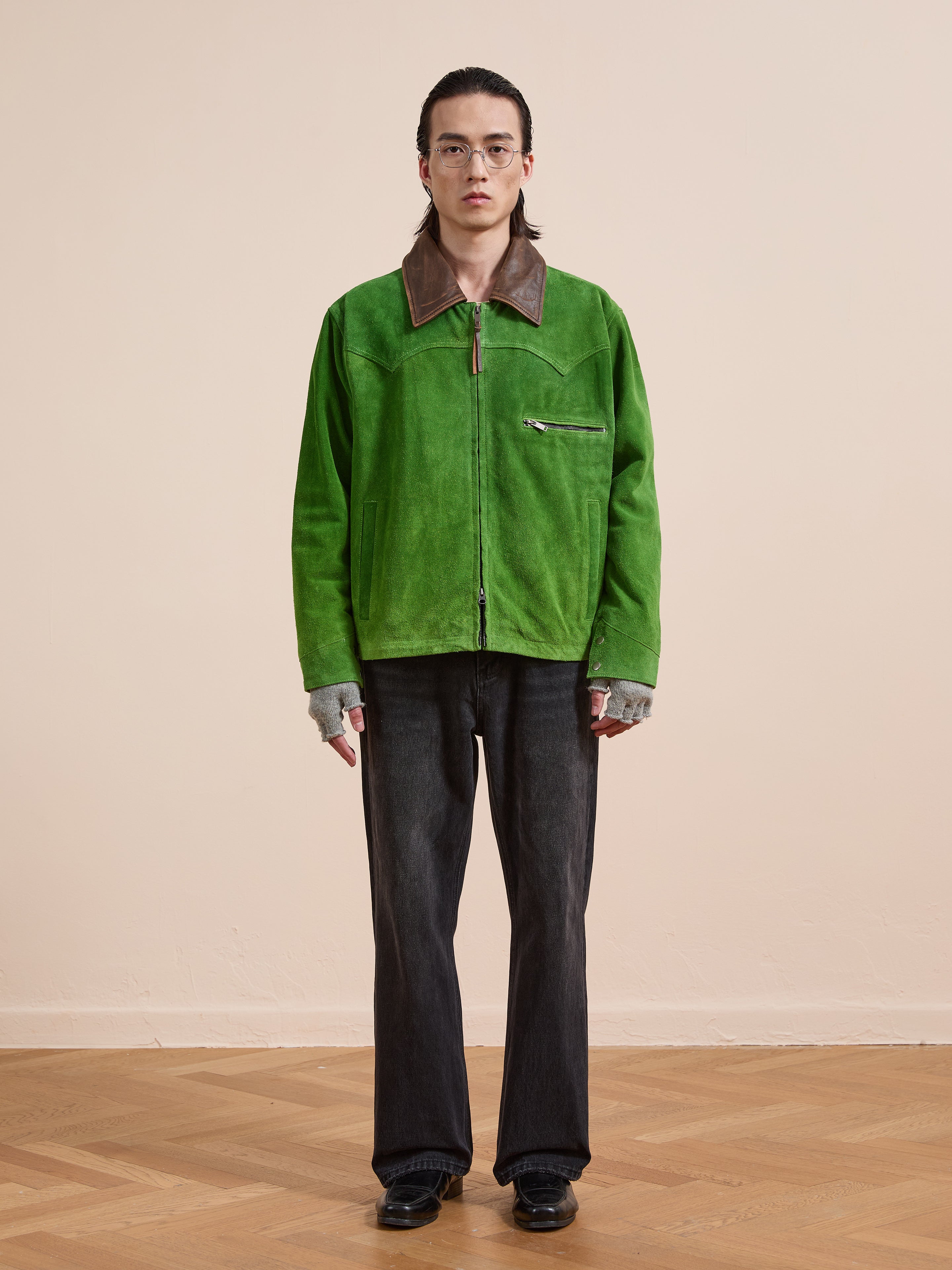 A person wearing the FOUND Suede Western Barn Jacket in bright green with a brown collar, gray fingerless gloves, dark jeans, and black shoes stands on a wooden floor against a plain beige wall.