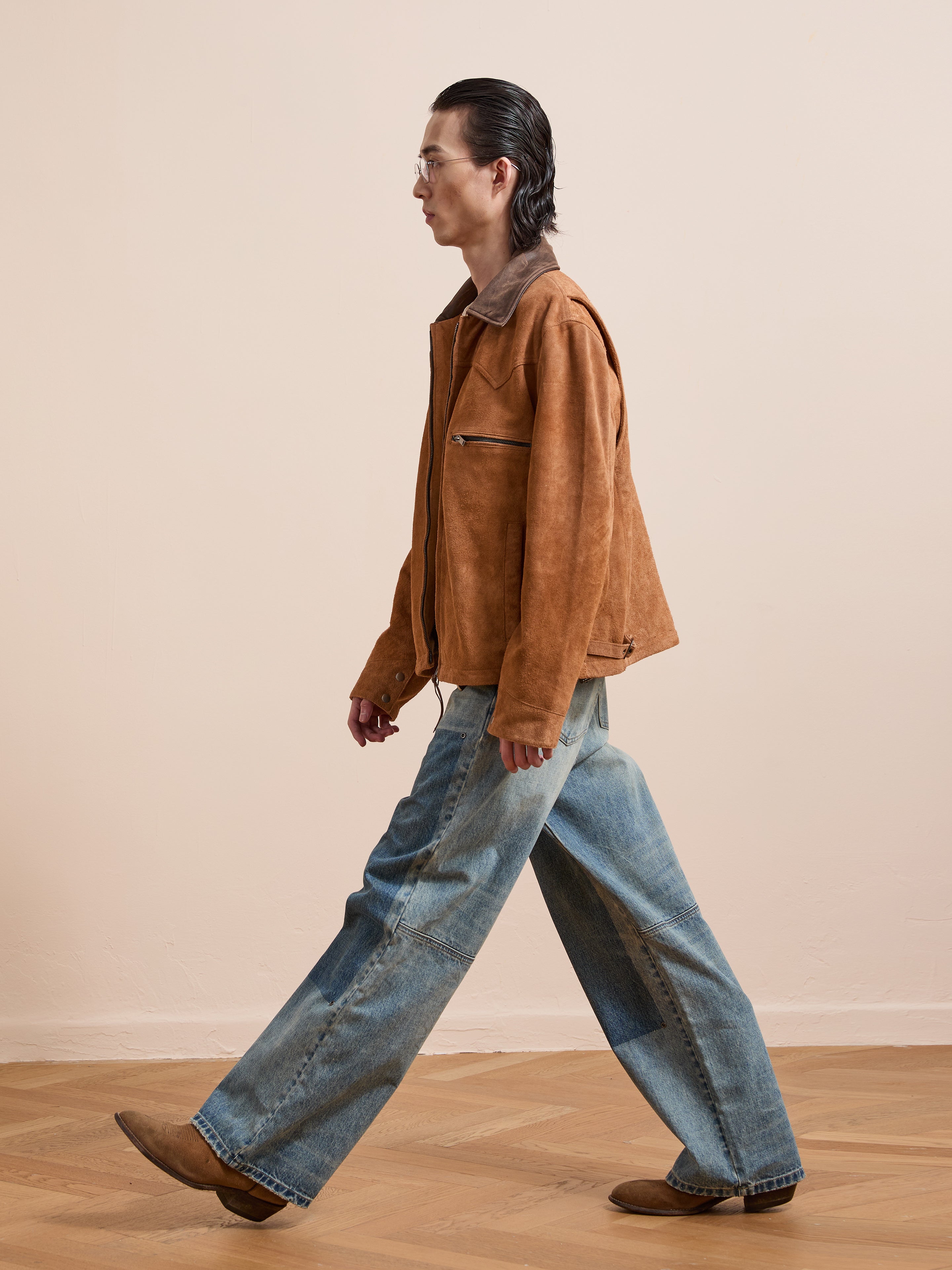 A person with slicked-back hair walks indoors on a wooden floor wearing the FOUND Suede Western Barn Jacket, wide-leg blue jeans, and brown shoes.