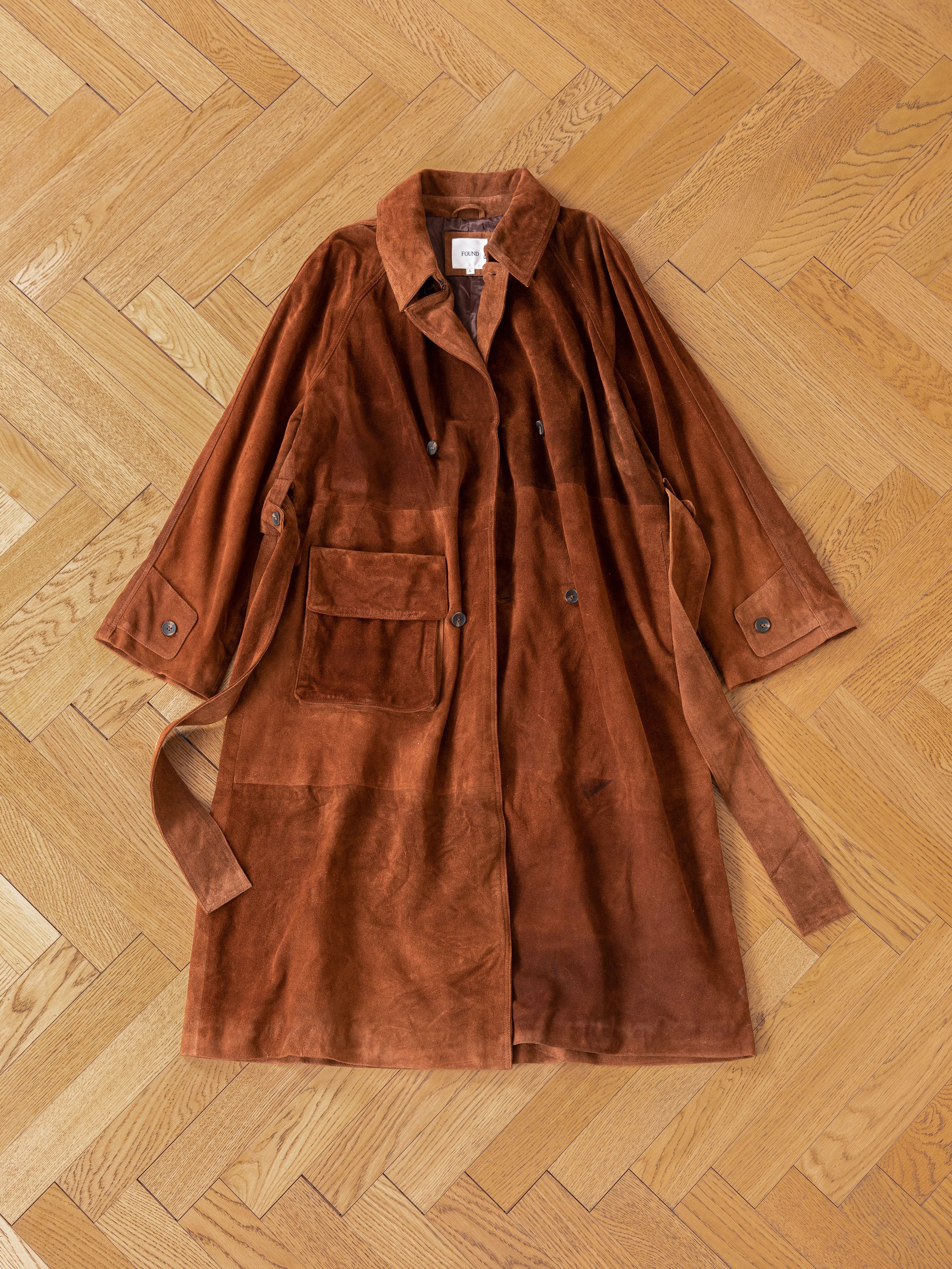 A unisex suede leather trench coat by FOUND, crafted ethically with a large front pocket, is spread out on a wooden herringbone floor.