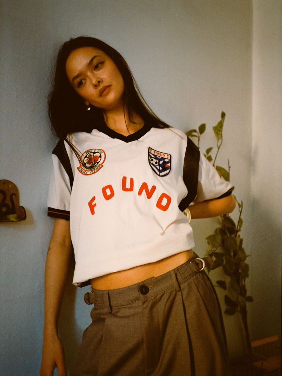 A person leans against a wall wearing a white Union Soccer Jersey by FOUND, paired with gray pants, exuding a contemporary edge. A plant provides a subtle backdrop, accentuating the stylish vibe.