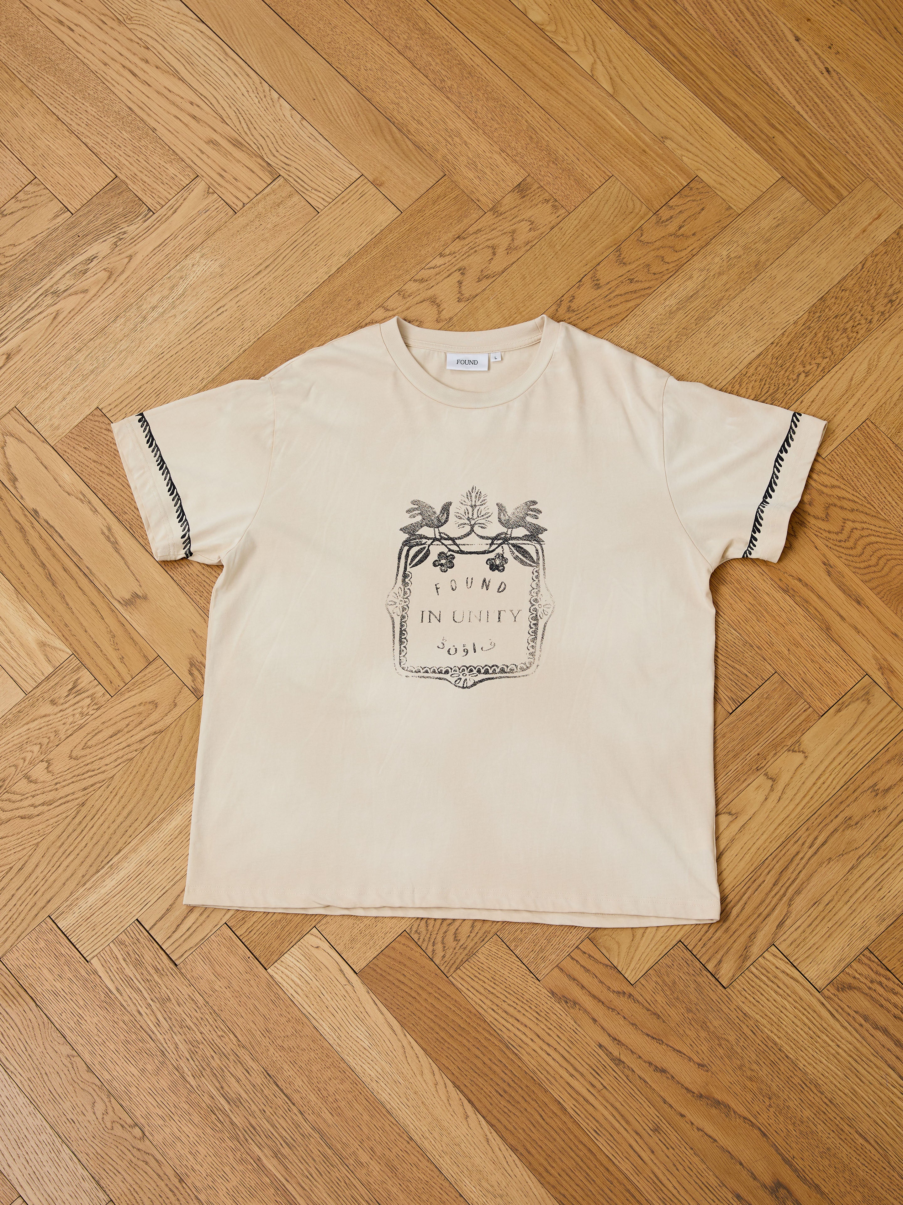 The FOUND Unity Crest Tee is a beige unisex t-shirt made of 100% cotton, featuring a black vine print and text on the front, displayed flat on a wooden parquet floor.
