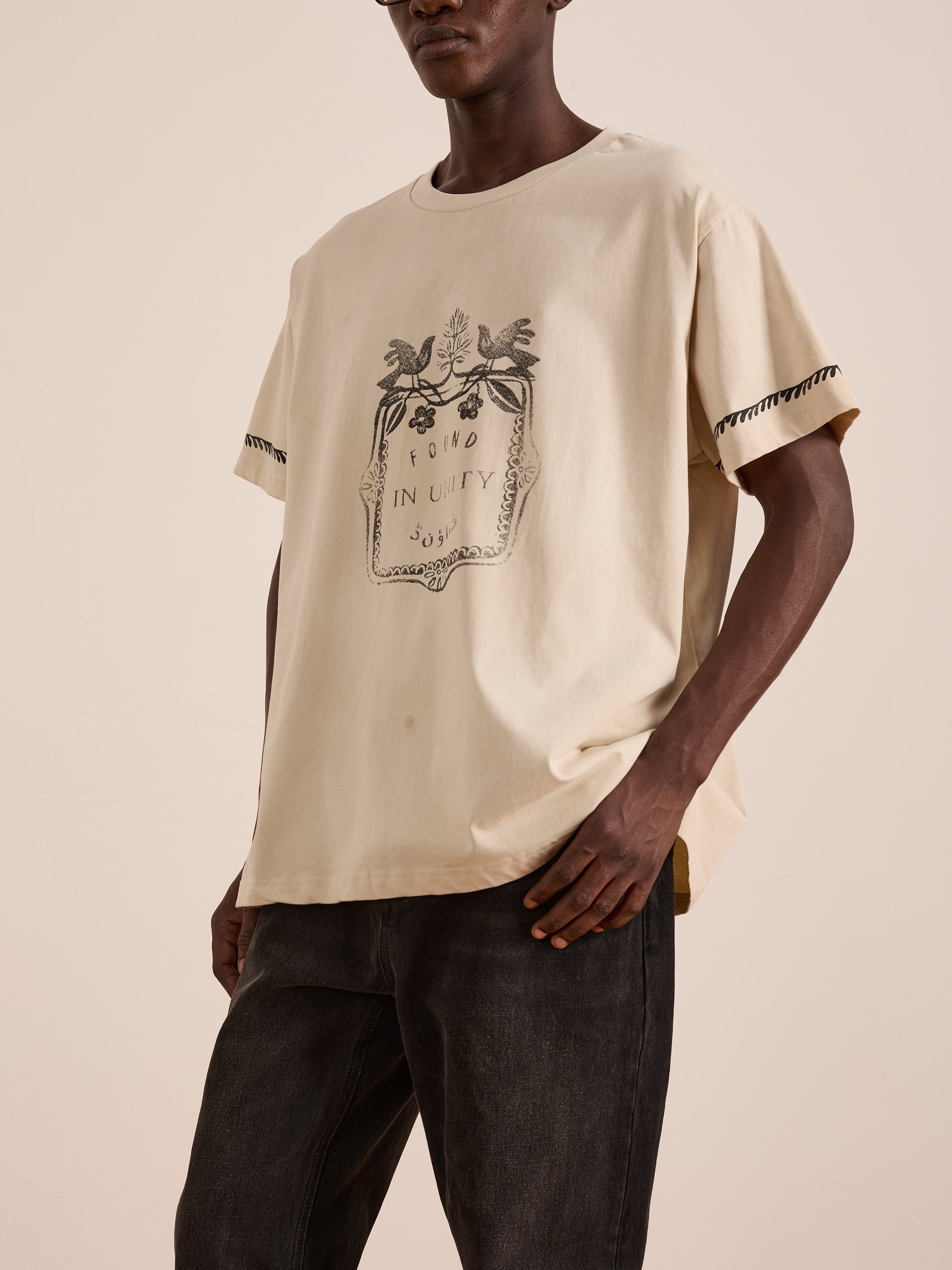 A person wearing the FOUND Unity Crest Tee, a loose mid-weight beige unisex cotton shirt with a decorative Urdu graphic, paired with dark pants and standing against a plain light background.