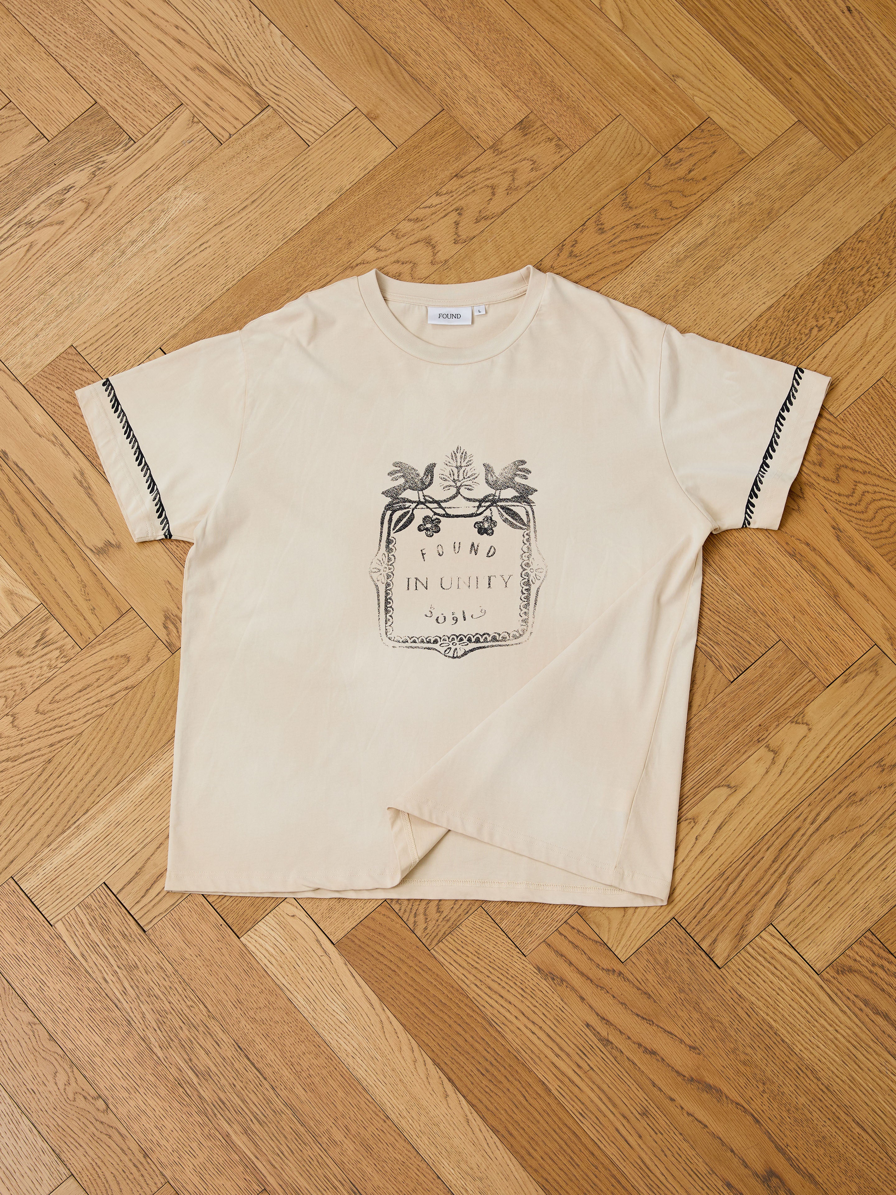 The Unity Crest Tee by FOUND is a beige unisex mid-weight cotton shirt featuring a black leaf and flower graphic with "FOUND IN UNITY SOSSIS" on the front, shown laid flat on a wooden parquet floor.