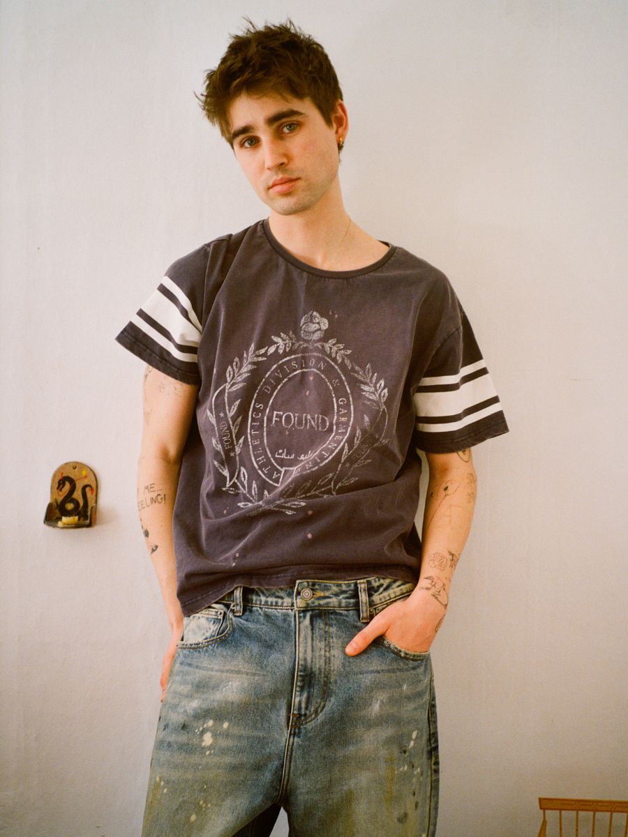 A person with short hair, hands in pockets, stands against a wall wearing the FOUND Collegiate Crest Tee and jeans. The unisex relaxed fit tee enhances the vintage athletic vibe, effortlessly showcasing their tattoos.