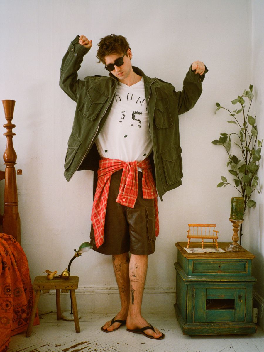 In a room filled with eclectic furniture and decor, someone channels retro appeal wearing sunglasses, a green jacket, and a white printed shirt. A red plaid shirt is tied at the waist over shorts and sandals, completing their vintage sportswear look with FOUND's Distressed LS Team Jersey.