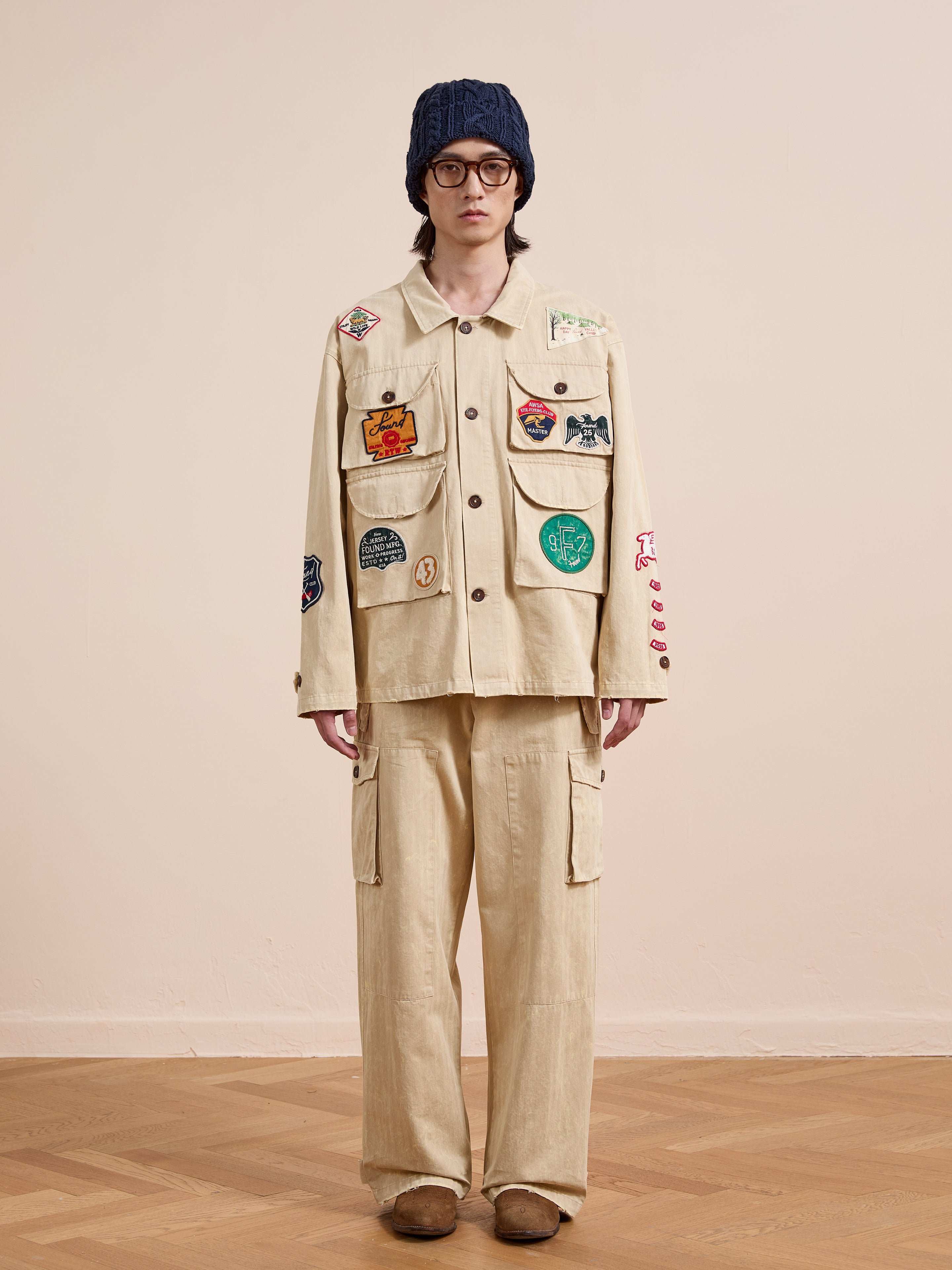 A person in glasses, a blue knit hat, and the FOUND Herringbone Utility Patchwork Jacket stands on a wooden floor against a plain, light-colored wall.