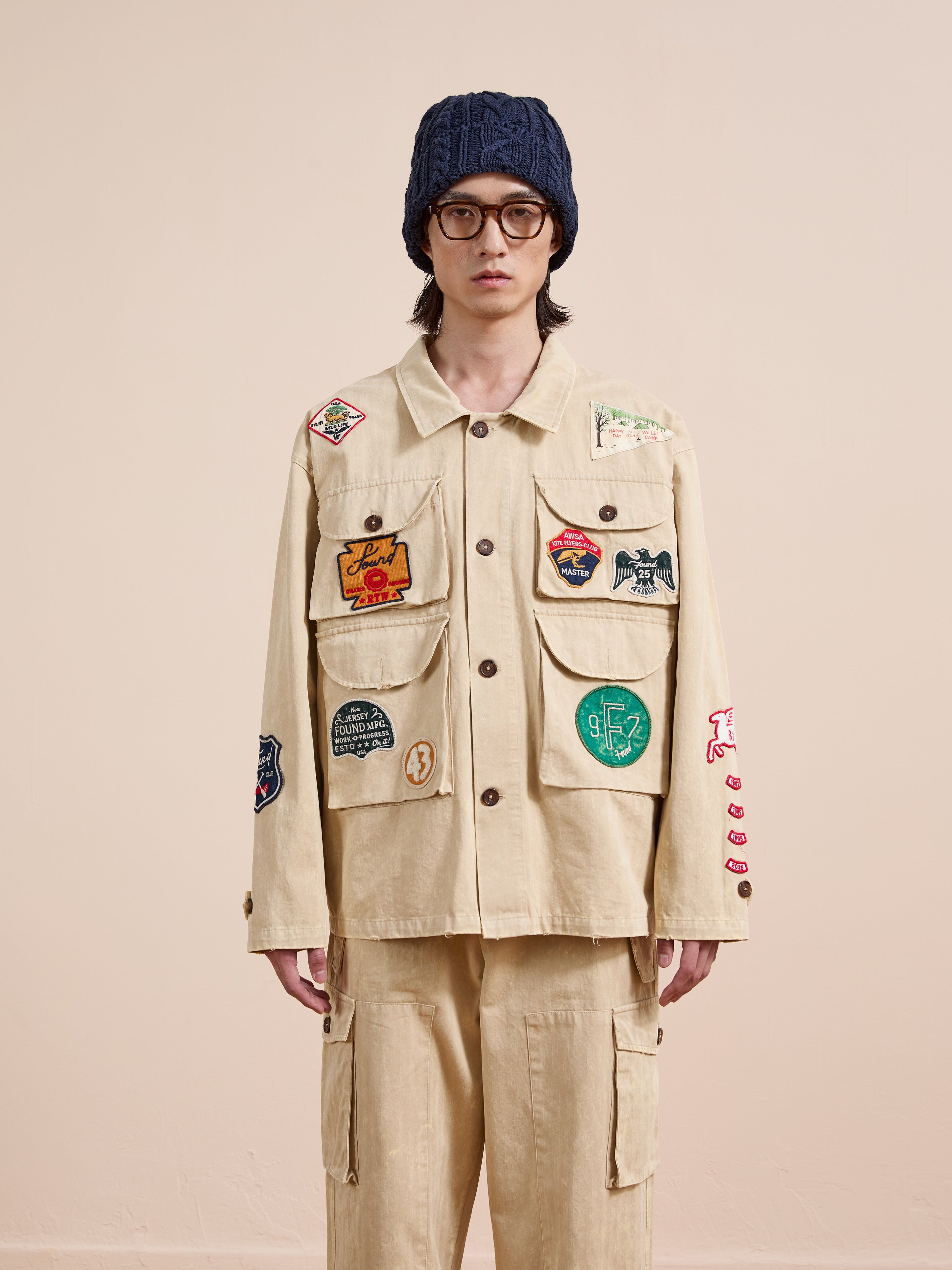 Wearing glasses, a knitted beanie, and FOUND’s Herringbone Utility Patchwork Jacket with pants, a person stands against a plain light background.