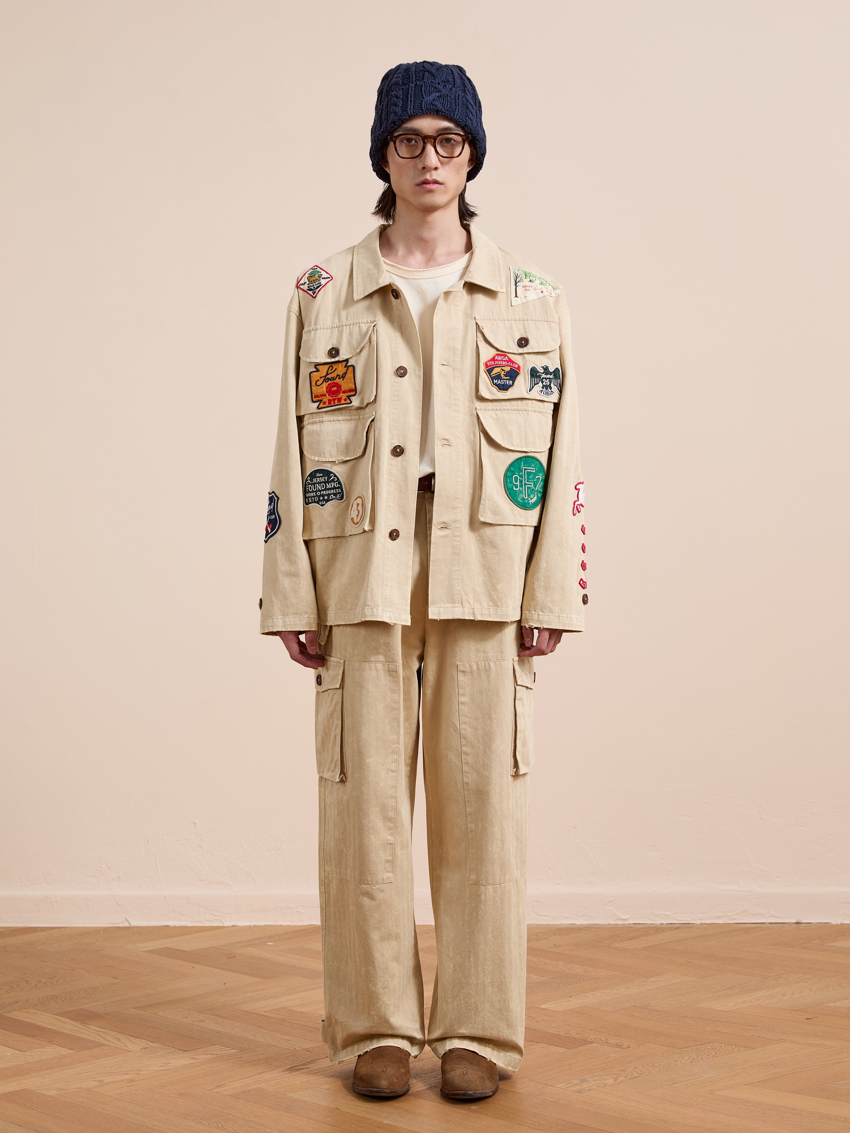 Person standing indoors against a neutral background, wearing FOUND's Herringbone Utility Patchwork Jacket and matching pants, styled with a navy knit hat, round glasses, and brown shoes.