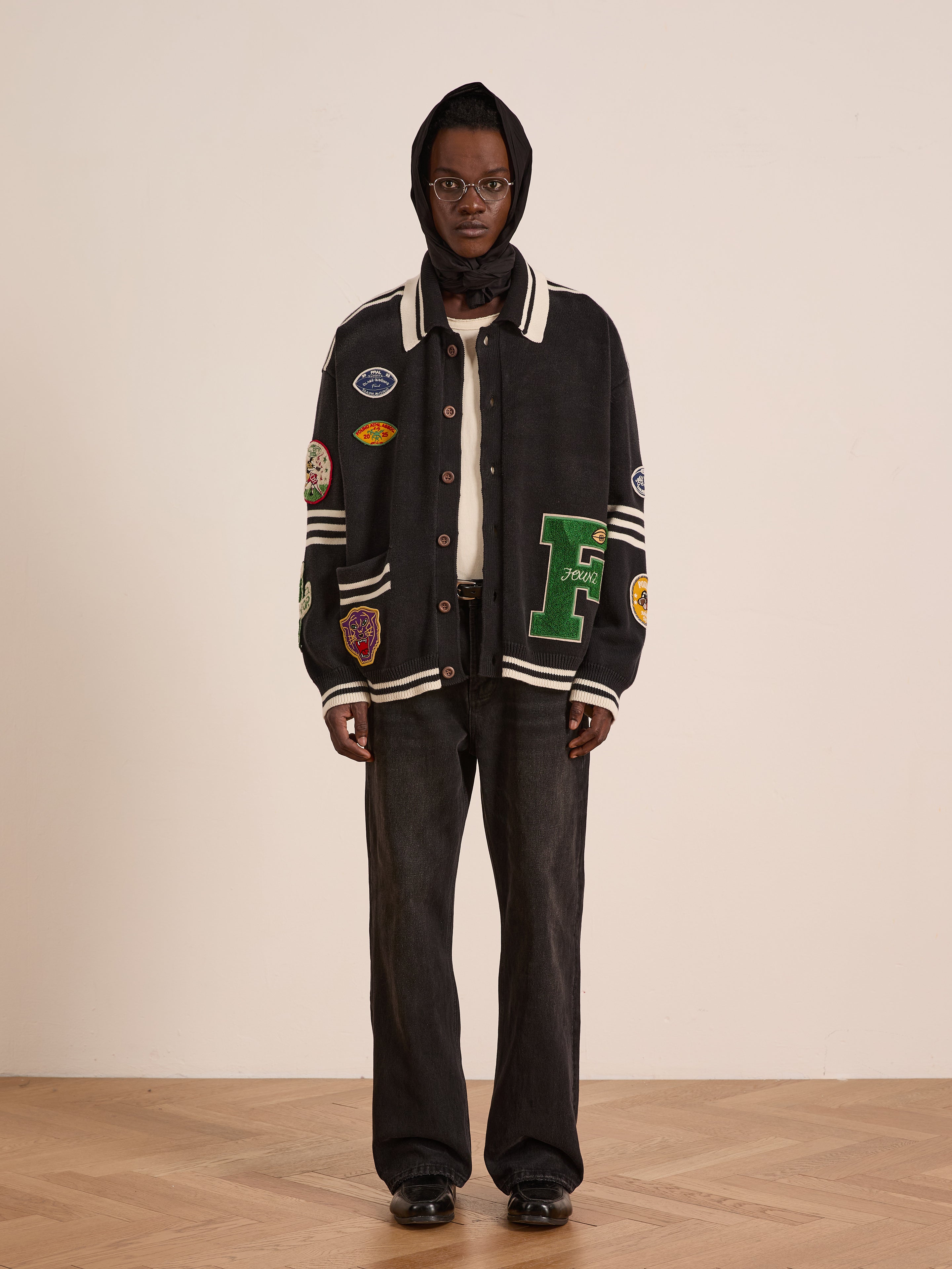 A person stands indoors against a plain light-colored wall, wearing a dark hooded scarf, FOUND Varsity Patch Collared Cardigan with patches, wide-leg dark jeans, and black shoes.