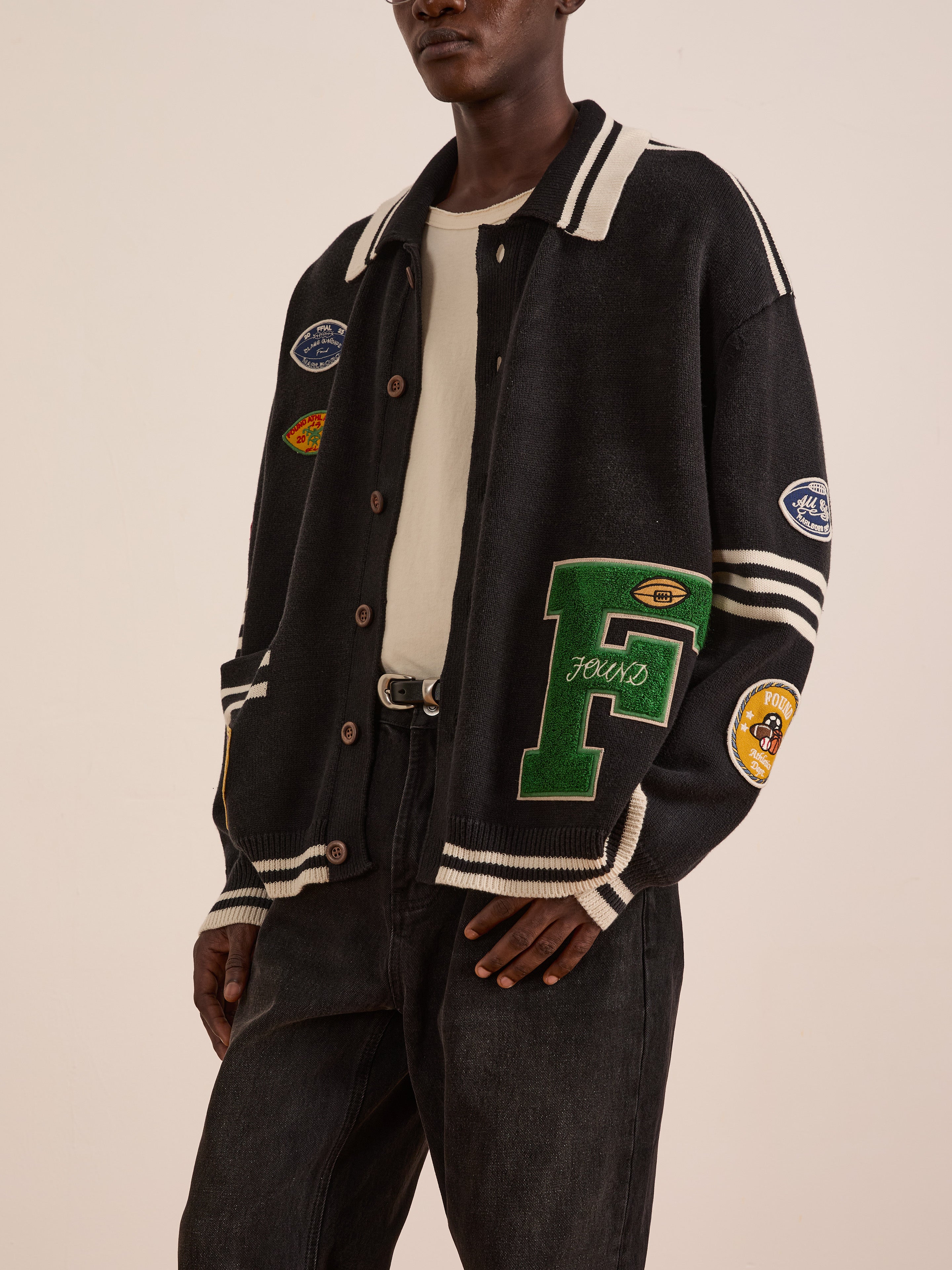 A person wears the FOUND Varsity Patch Collared Cardigan with varsity-inspired patches over a white shirt and black jeans, standing against a plain background.