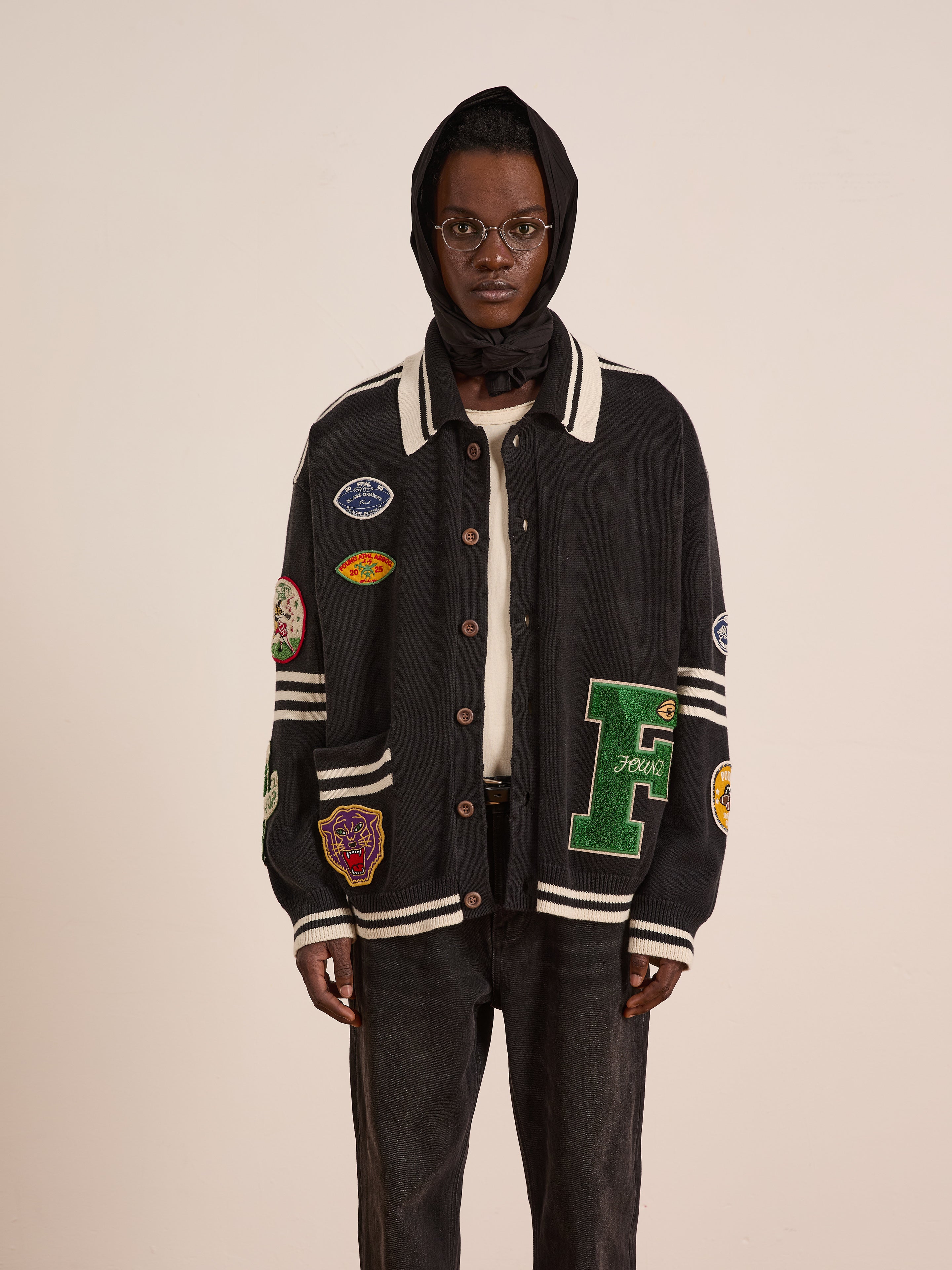 A person in glasses and a black hood faces forward, wearing the FOUND Varsity Patch Collared Cardigan with patches and dark jeans against a plain background.