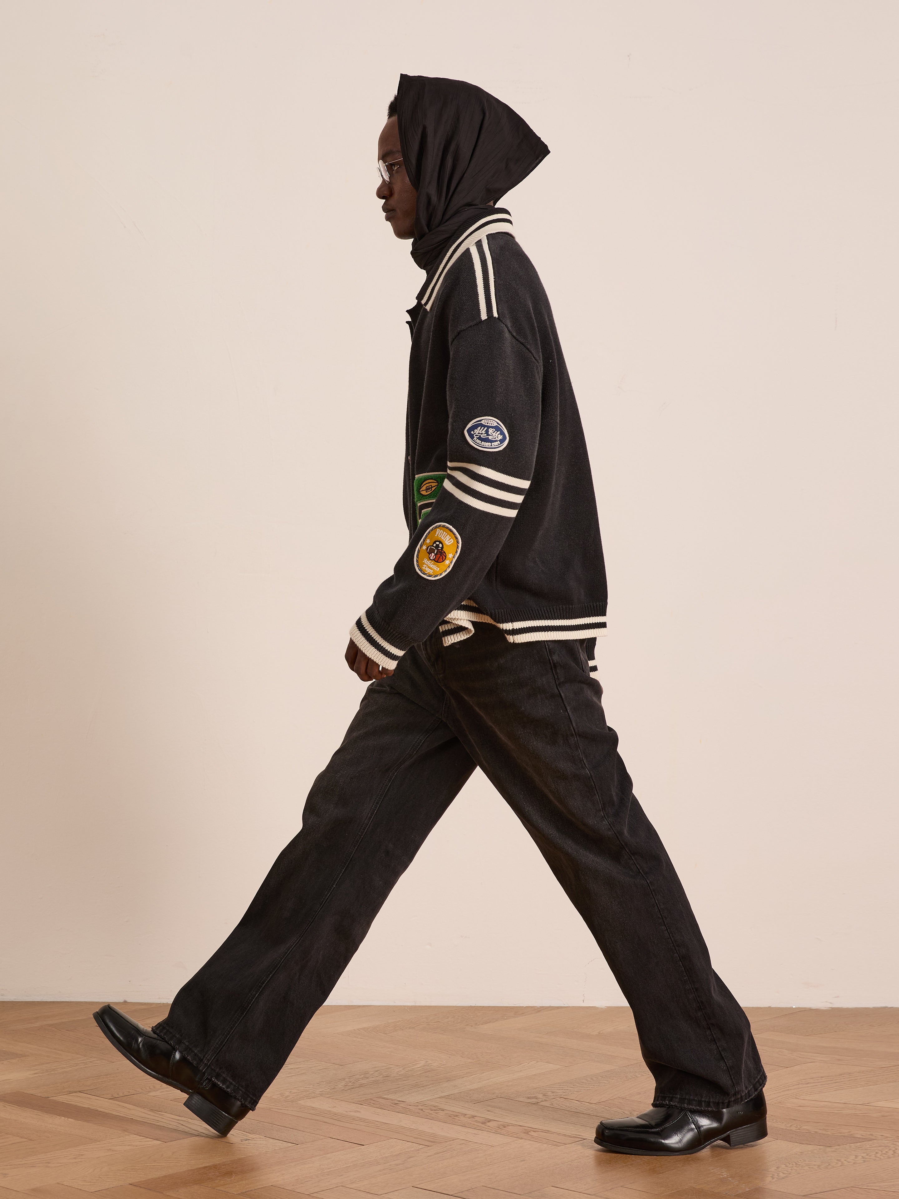 A person wearing a black hood, FOUND Varsity Patch Collared Cardigan, black pants, and black shoes walks indoors on a wooden floor.