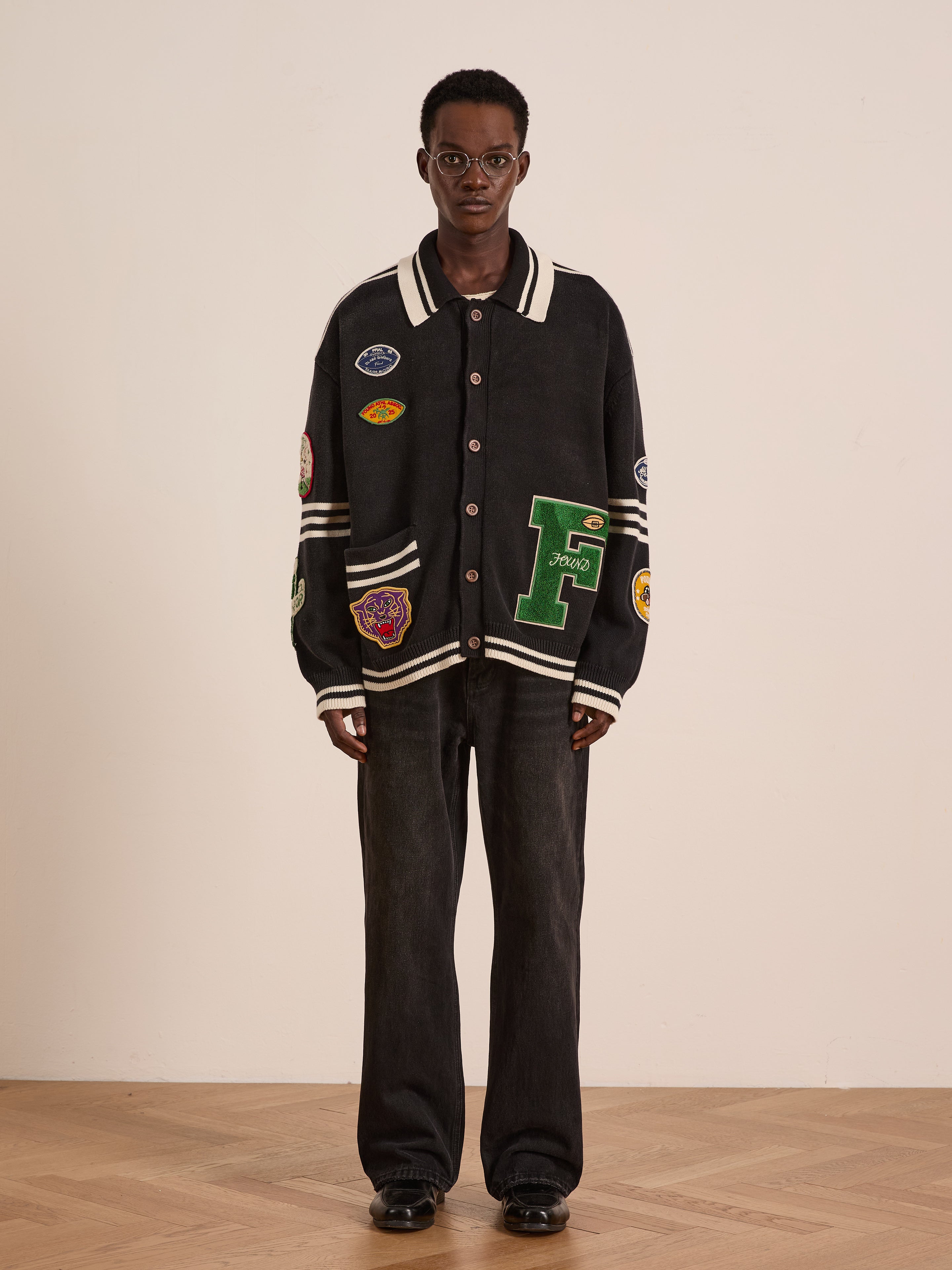 A person stands indoors wearing glasses, a FOUND Varsity Patch Collared Cardigan, black jeans, and black shoes against a plain background.