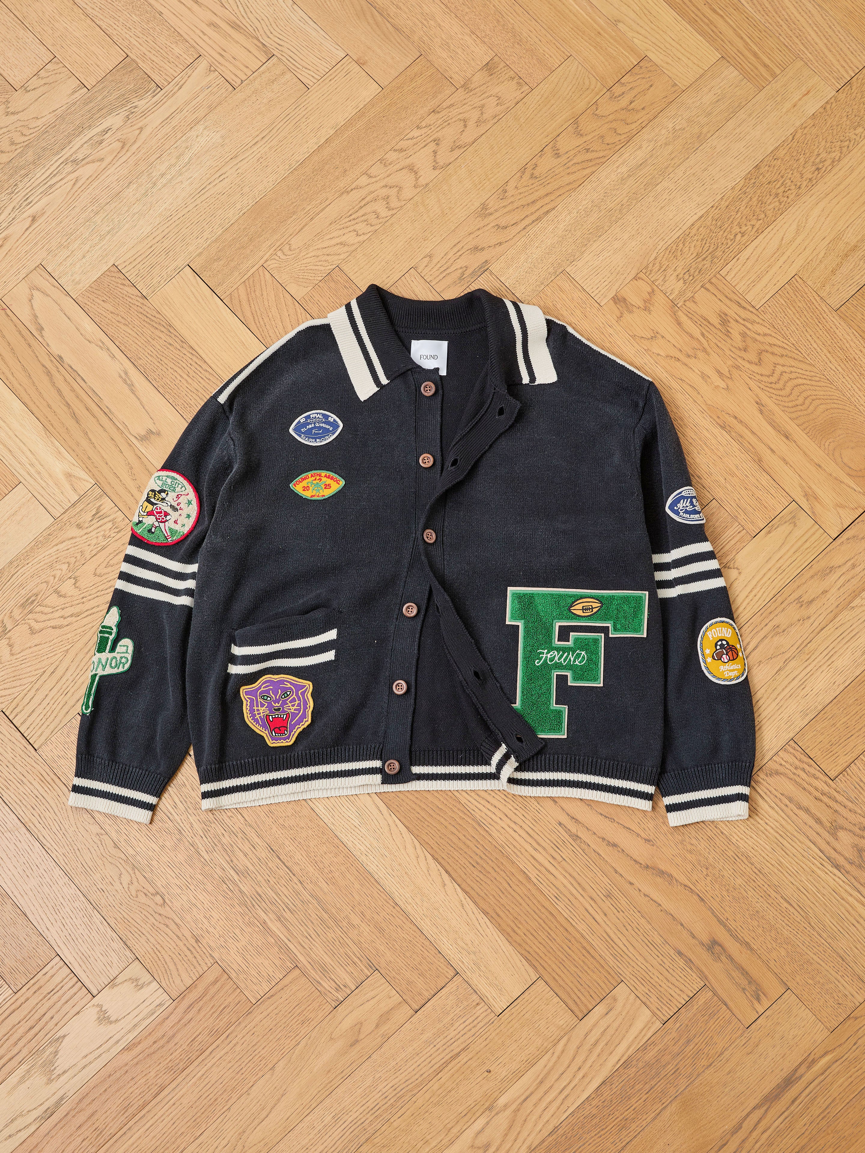 The FOUND Varsity Patch Collared Cardigan, featuring black fabric, white stripes, button closure, and colorful chenille patches, is laid flat on a wooden floor.