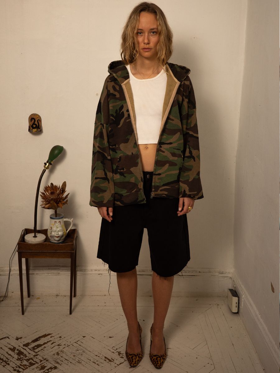A person with shoulder-length hair stands indoors wearing a FOUND Washed Camouflage Zip Hoodie, white cropped top, black shorts, and leopard print shoes. Behind them, a small table with a lamp and decor complements the unisex ensemble in this stylish setting.
