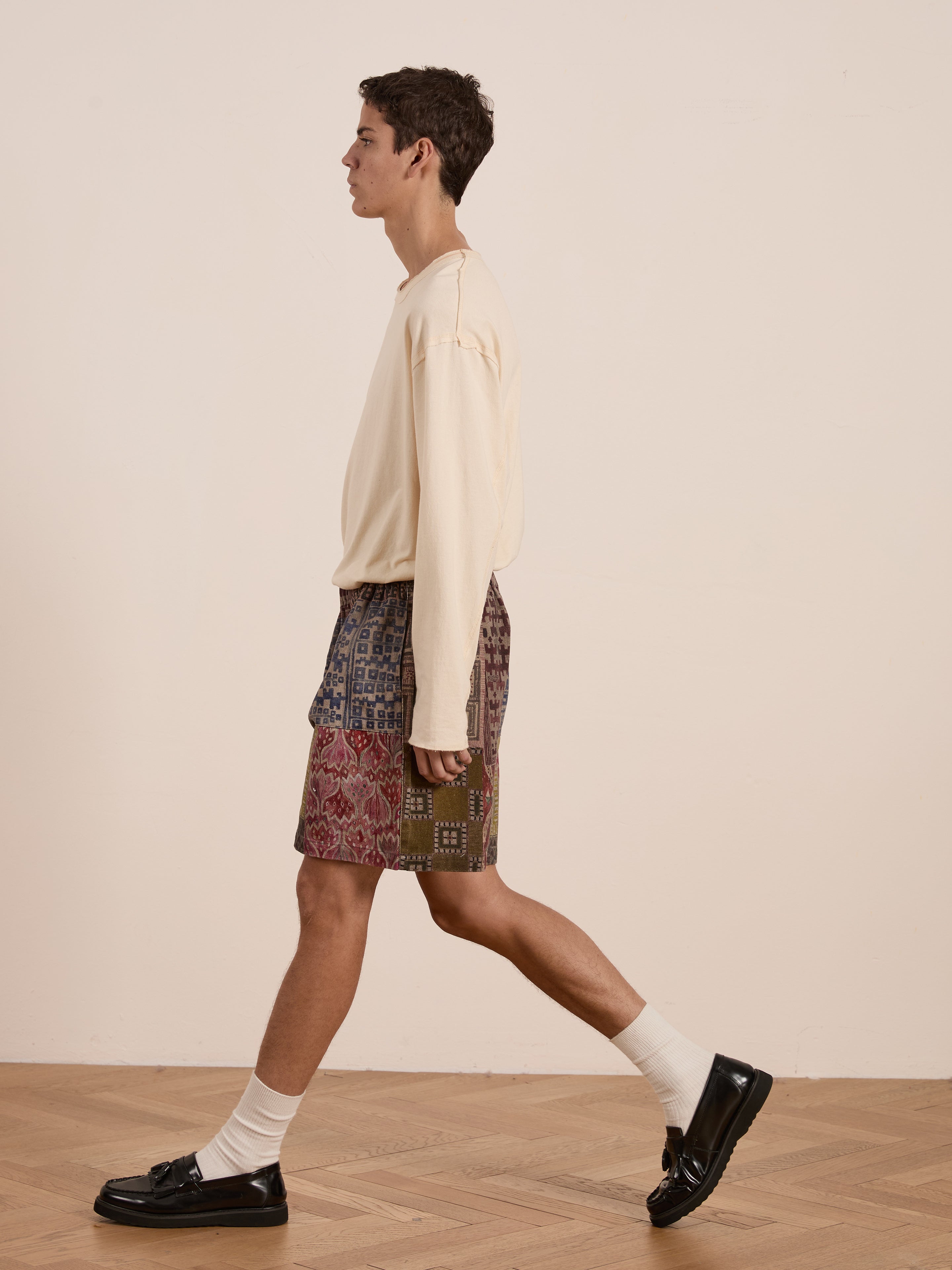 Someone in a long-sleeve beige shirt and FOUND's Watercolor Mosaic Work Shorts, featuring vintage-inspired patterns, walks confidently across a wooden floor, complementing the unisex relaxed fit with white socks and black loafers.