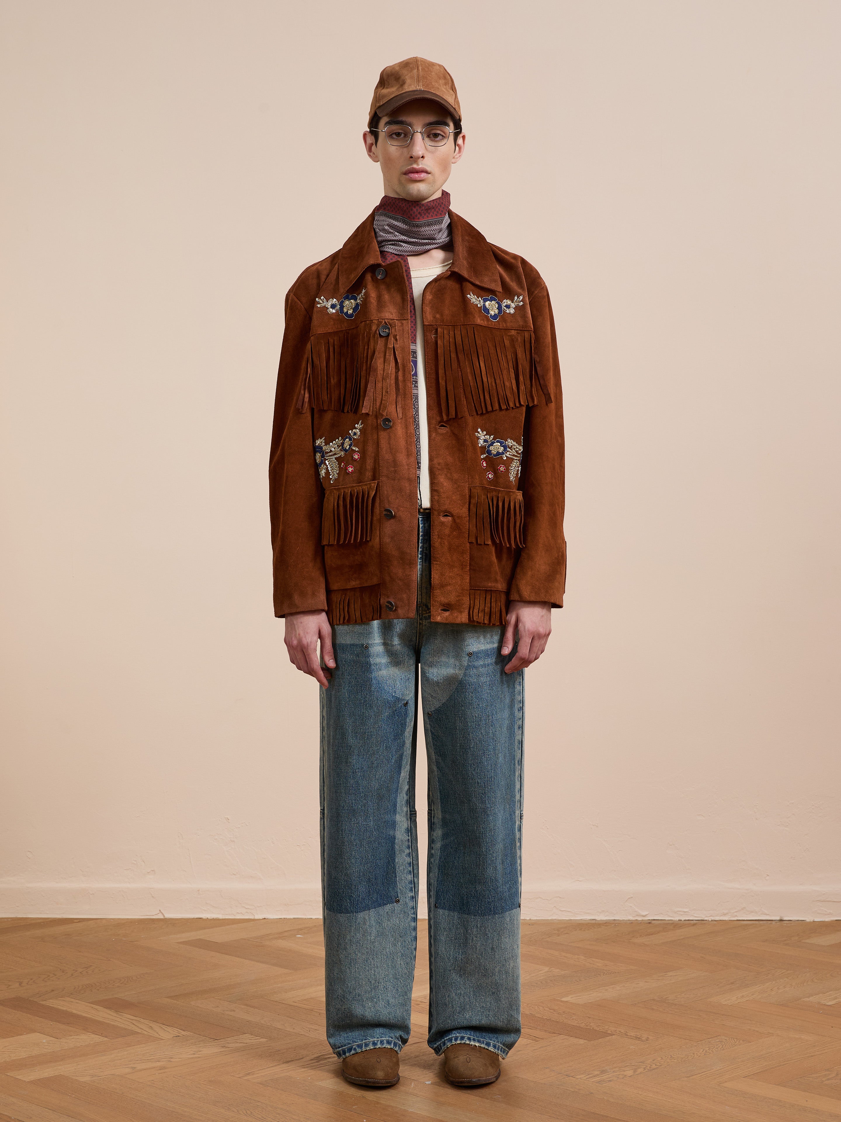 A person faces forward, wearing the FOUND Hand Embroidered Suede Fringe Jacket, blue patchwork jeans, brown shoes, a tan cap, and glasses against a plain background.