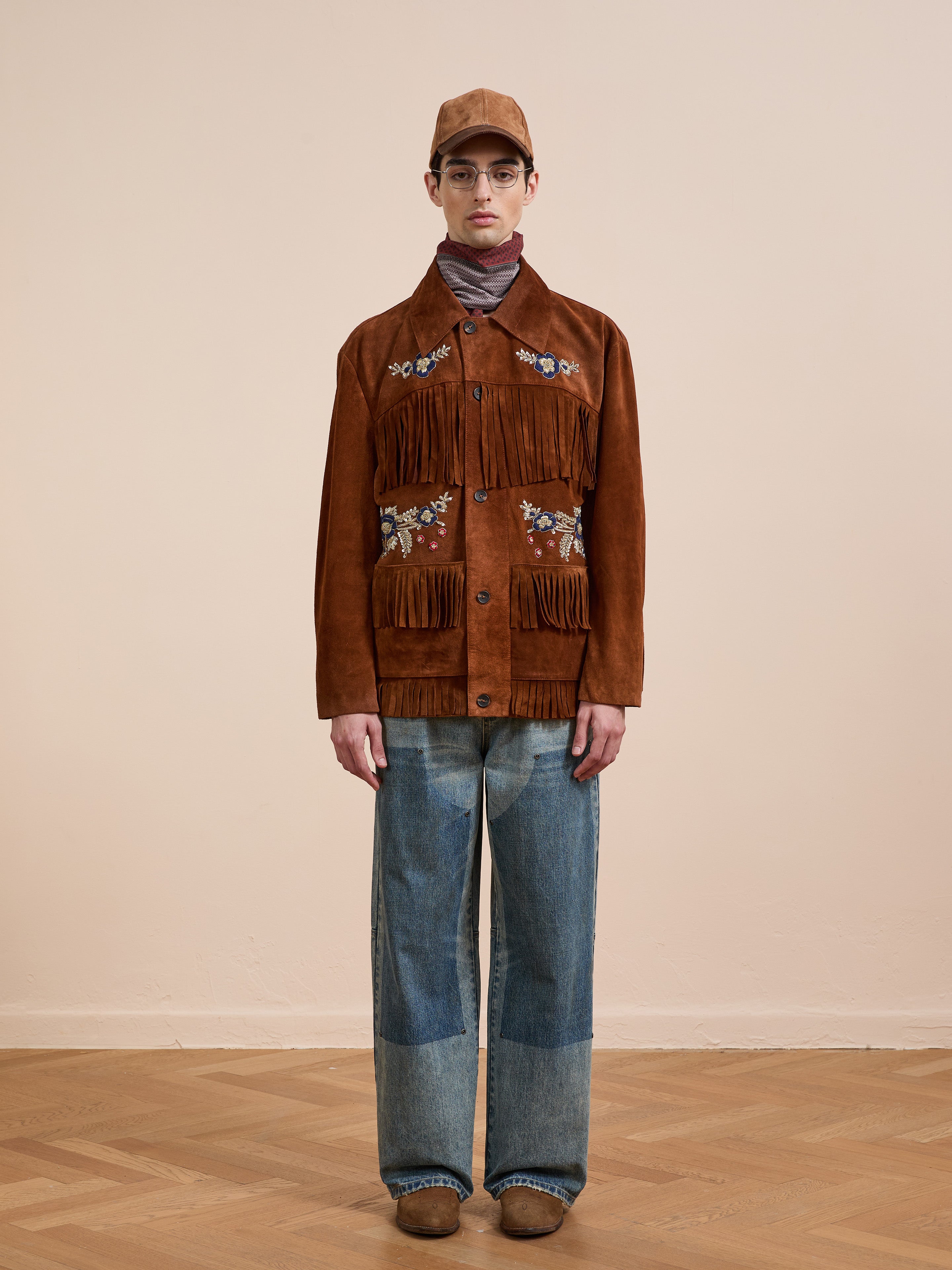 Indoors against a beige wall, a person wears the FOUND Hand Embroidered Suede Fringe Jacket, paired with blue jeans, brown shoes, a brown cap, and glasses.