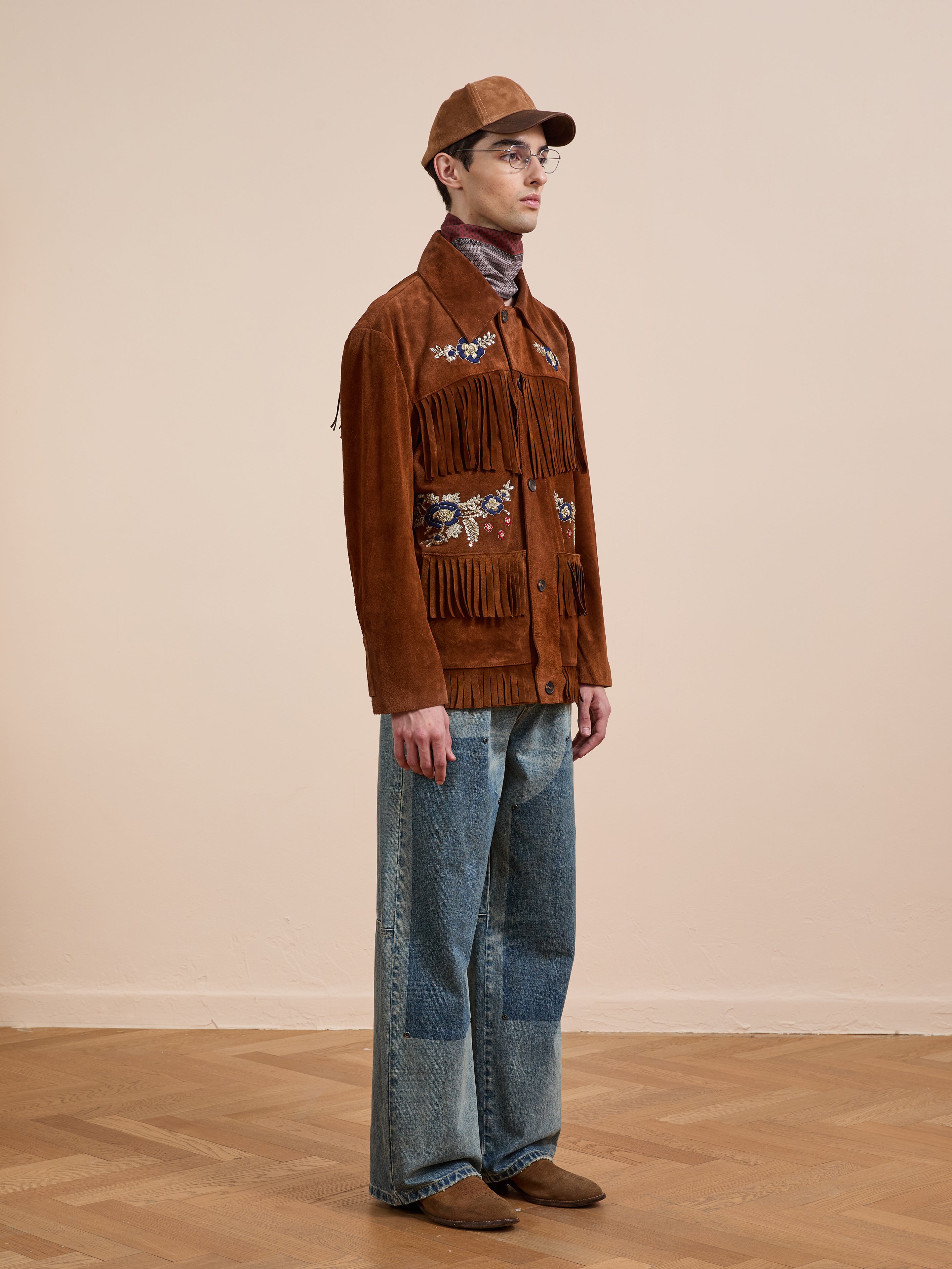 Indoors against a beige wall and wooden floor, a person wears the FOUND Hand Embroidered Suede Fringe Jacket with embroidered designs, paired with a brown cap, wide-legged jeans, and brown shoes.