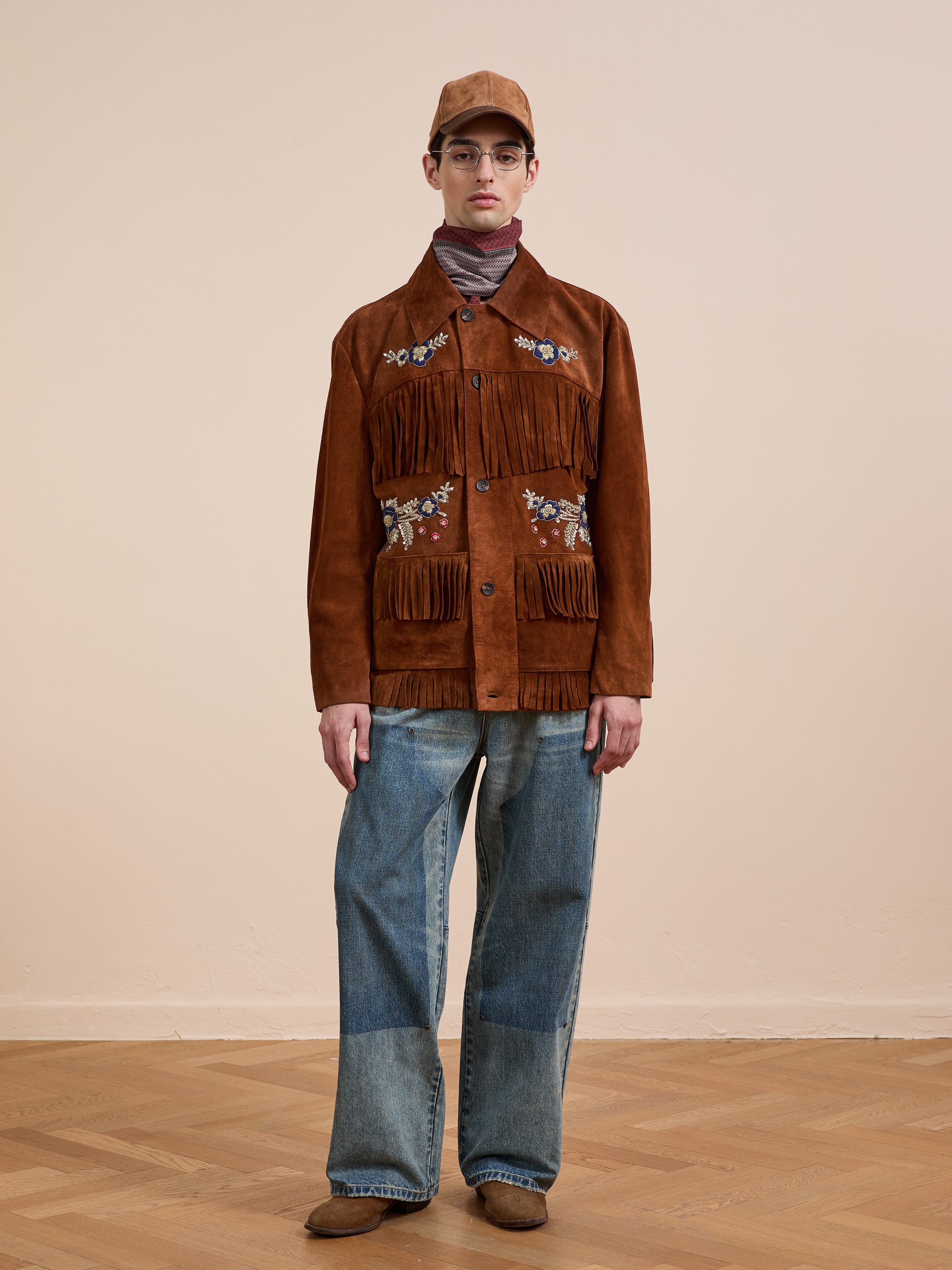 Indoors, a person wears the FOUND Hand Embroidered Suede Fringe Jacket with floral designs, paired with wide-leg blue jeans, a brown cap, and brown shoes.