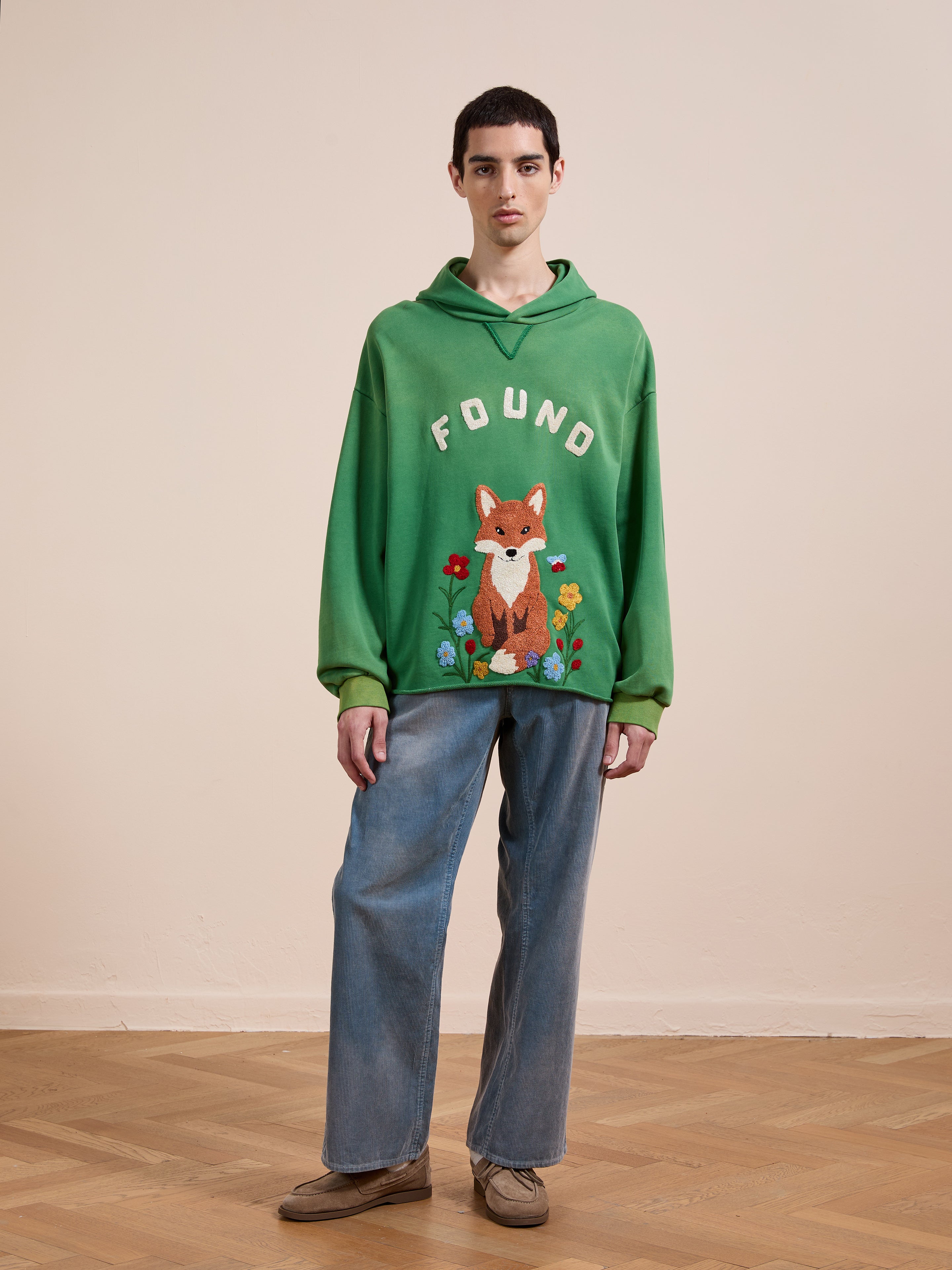Indoors, a person wears the FOUND Embroidered Wildlife Hoodie with a fox graphic and blue jeans, paired with tan shoes, standing on a wooden floor against a plain beige wall.