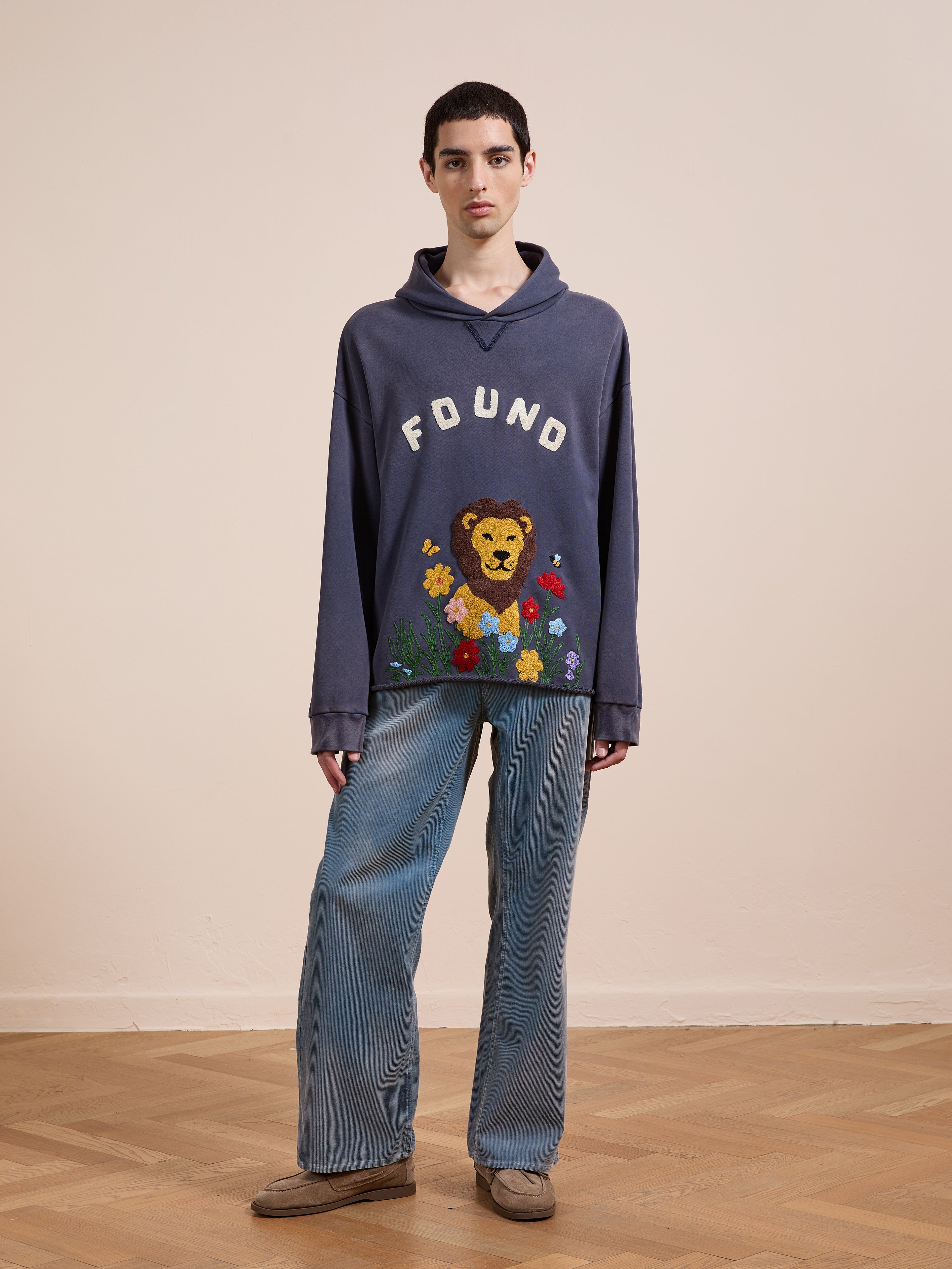 A person stands indoors wearing the FOUND Embroidered Wildlife Hoodie featuring a lion graphic and flowers, paired with loose blue jeans and brown shoes.
