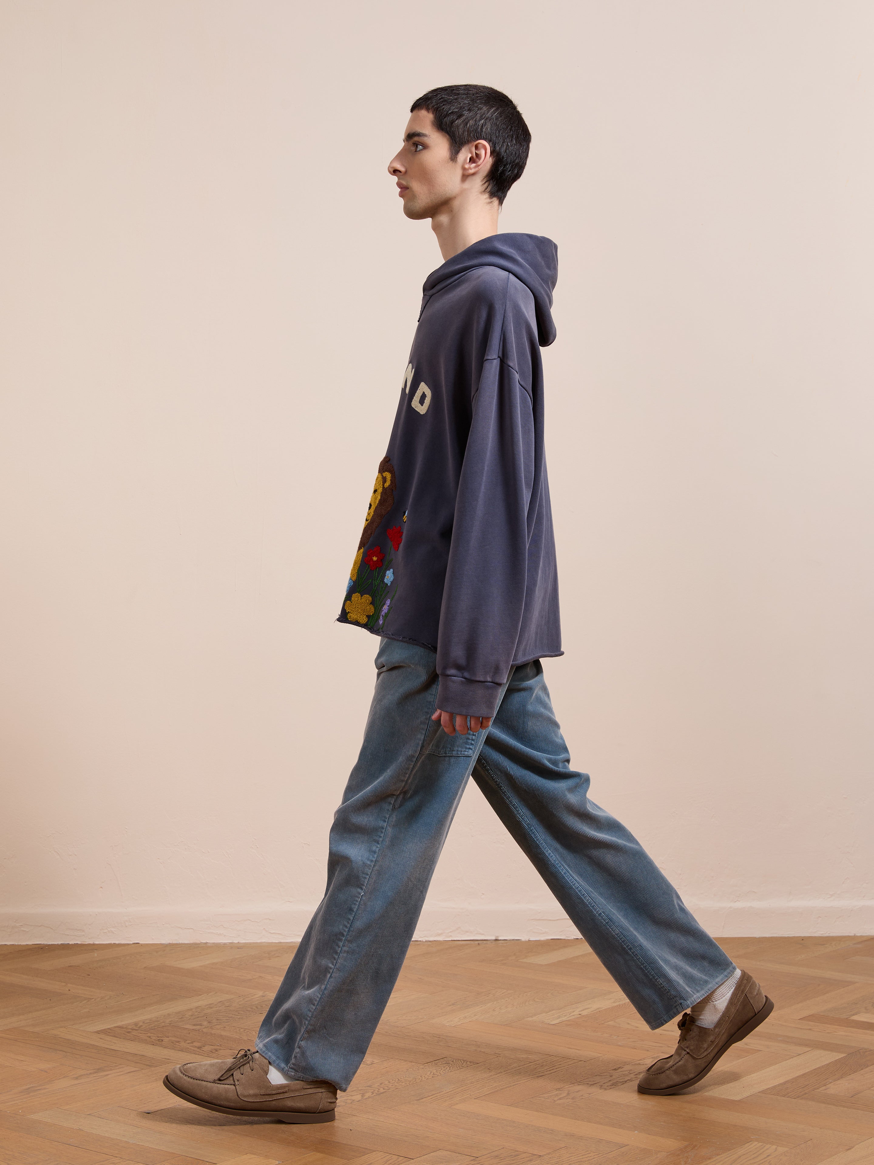 A young man in the FOUND Embroidered Wildlife Hoodie and loose jeans walks on a wooden floor, facing left in profile against a plain beige wall.